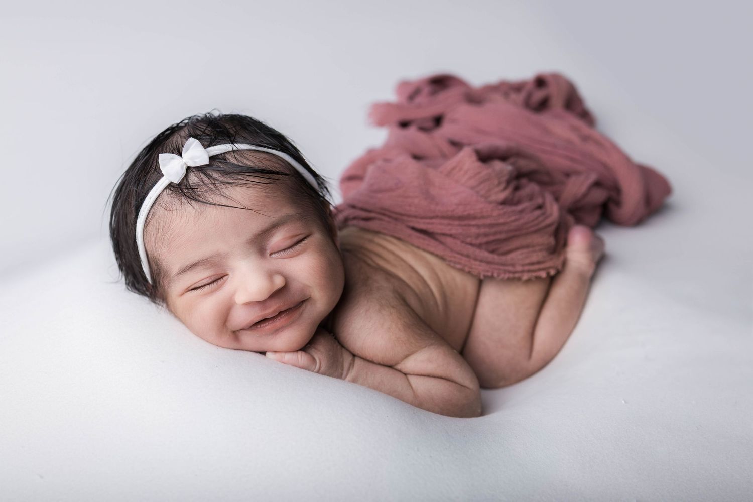 Smiling newborn baby girl wearing a white bow headband, wrapped in a dusty rose blanket on a white background.