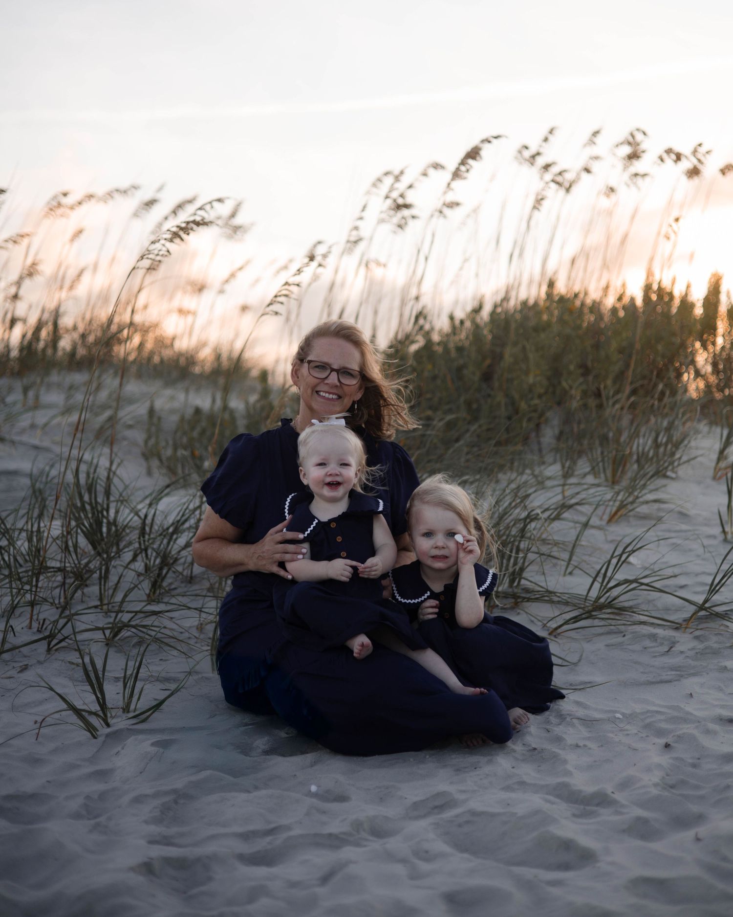 Family group poses together on beach sand with tall grass and sunset sky in background.