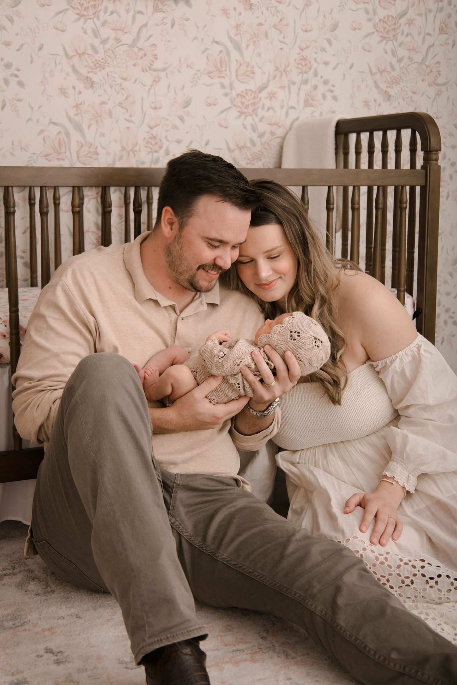 A couple sitting together on the floor by a wooden crib, tenderly gazing at their newborn baby in soft lighting.