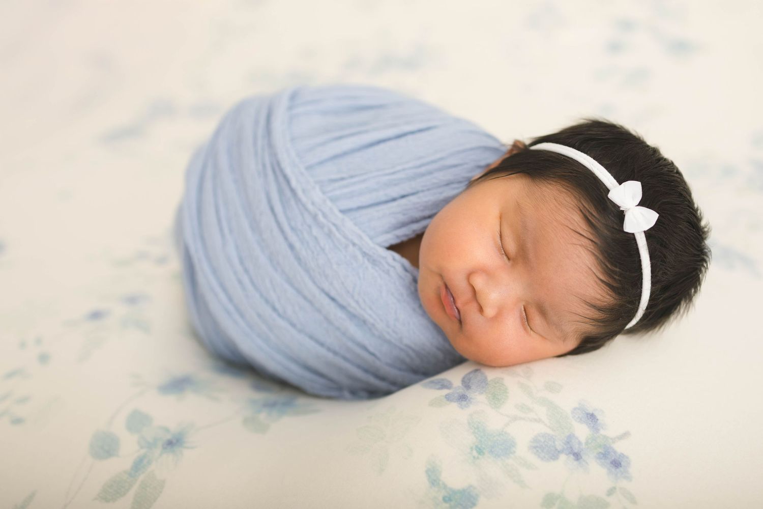 Sleeping newborn baby swaddled in blue wrap with white bow headband on floral background.
