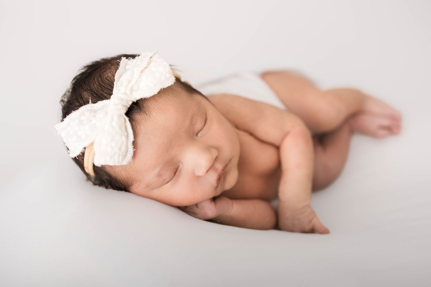 Peaceful newborn baby girl sleeping curled up, wearing a white lace bow headband, on a soft white background.