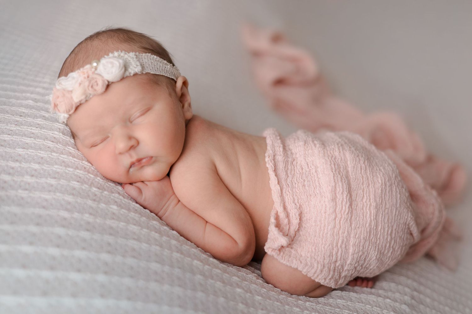 A sleeping newborn baby wrapped in soft pink fabric with a delicate lace headband rests on white textured bedding.