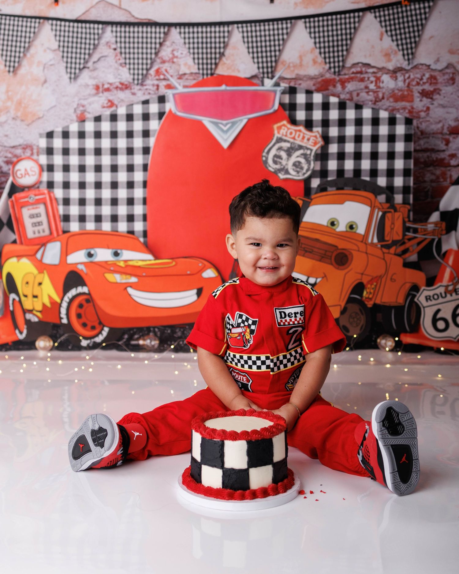 Toddler boy in red Cars outfit smiles beside checkered cake at a Disney Cars themed first birthday photo shoot.