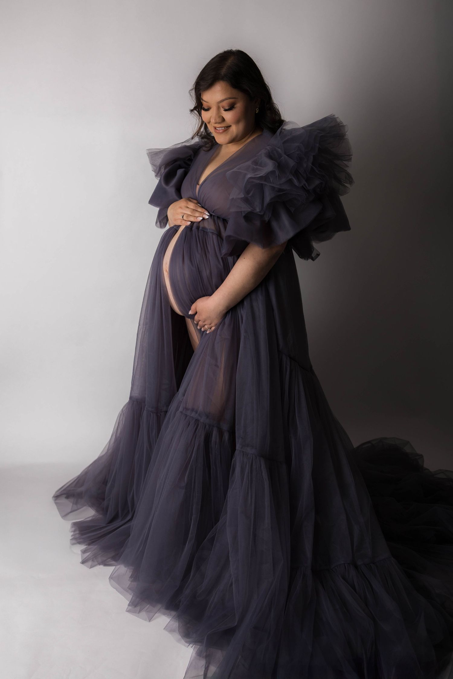 Pregnant woman in elegant dark purple tulle maternity gown with ruffled sleeves, posing gracefully in a studio.