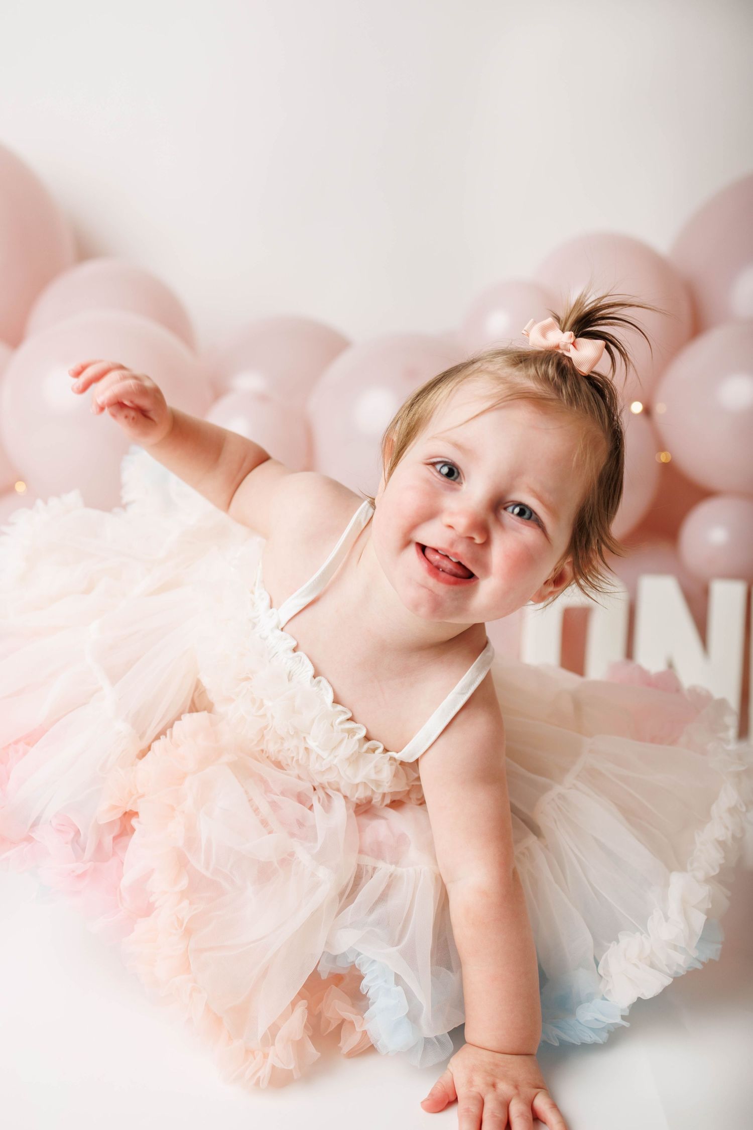Smiling toddler girl in pink tutu dress celebrating first birthday with pink balloons and ONE sign backdrop.