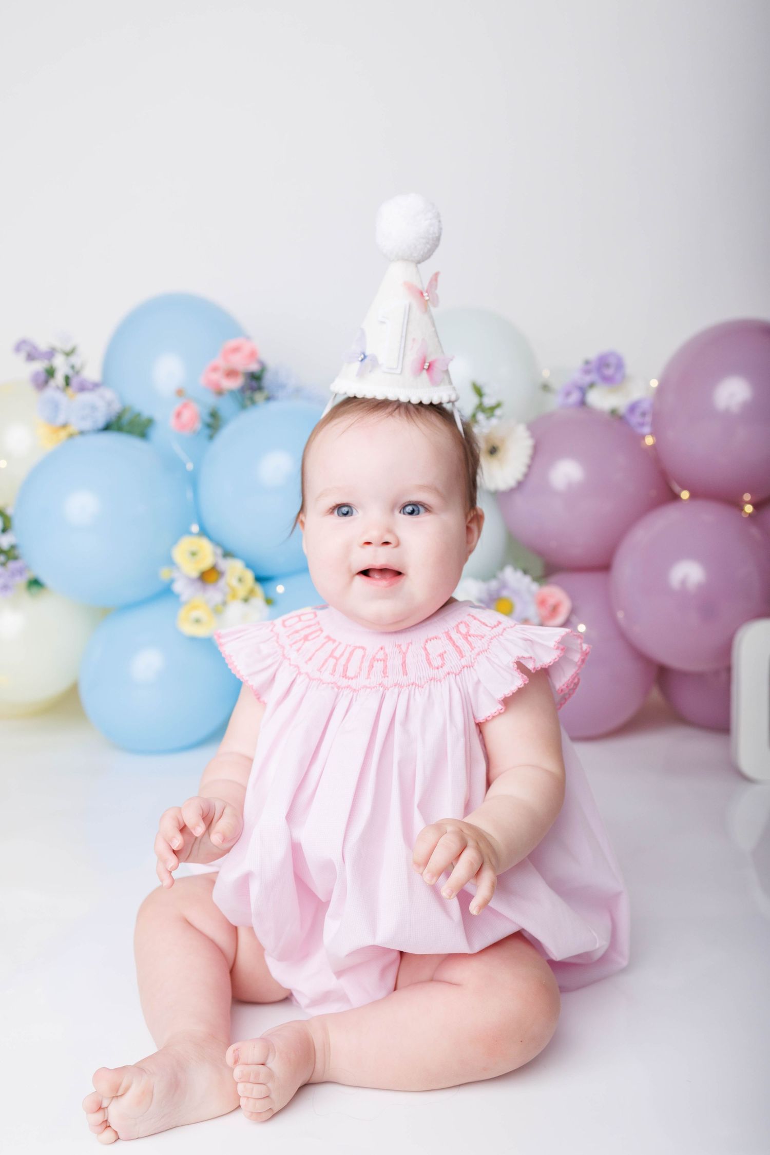 Baby girl in pink birthday dress and party hat sits surrounded by colorful pastel balloons at her first birthday celebration.