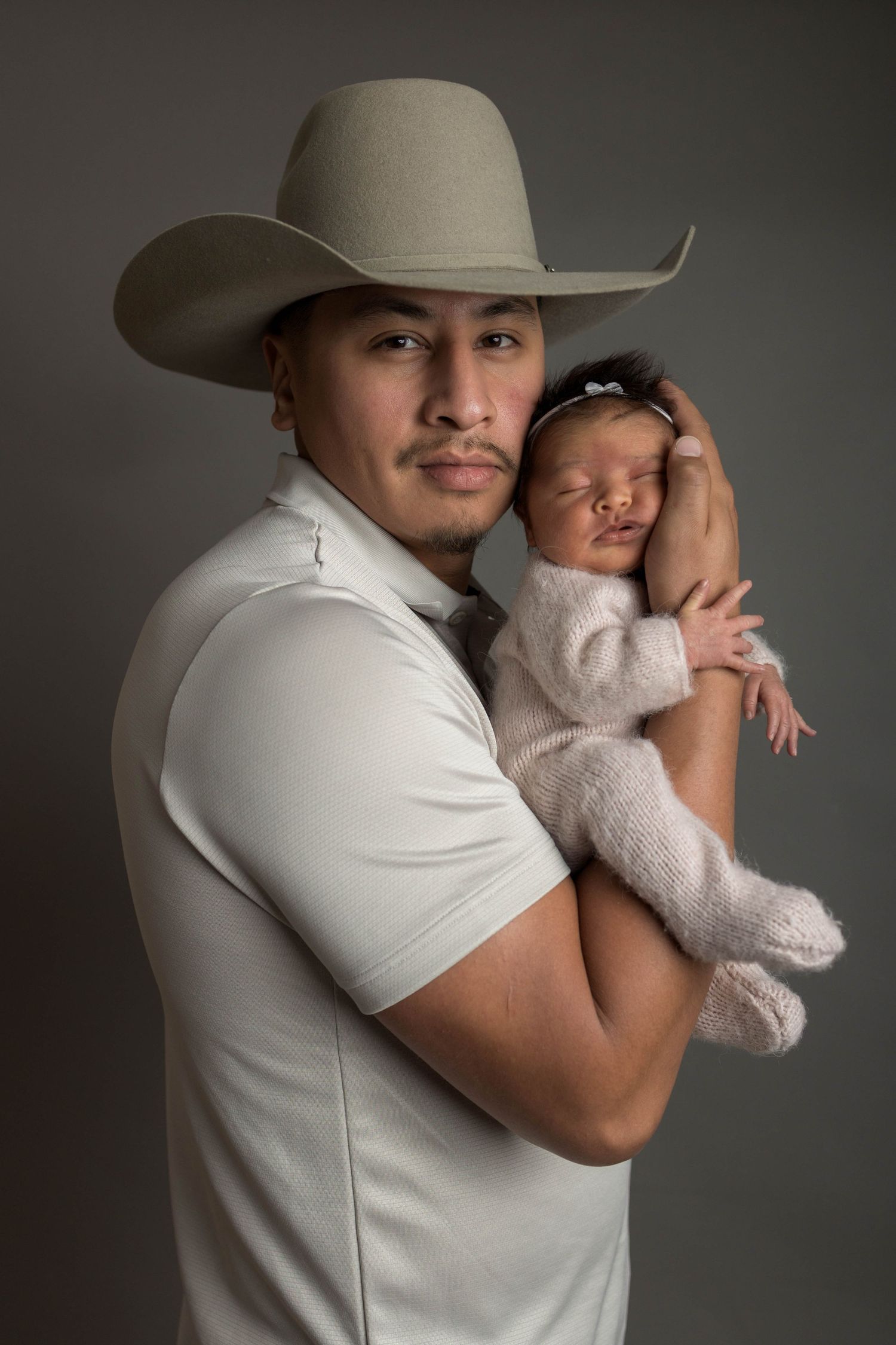 Man in cowboy hat tenderly holds sleeping newborn baby in white knit outfit for studio portrait photo.