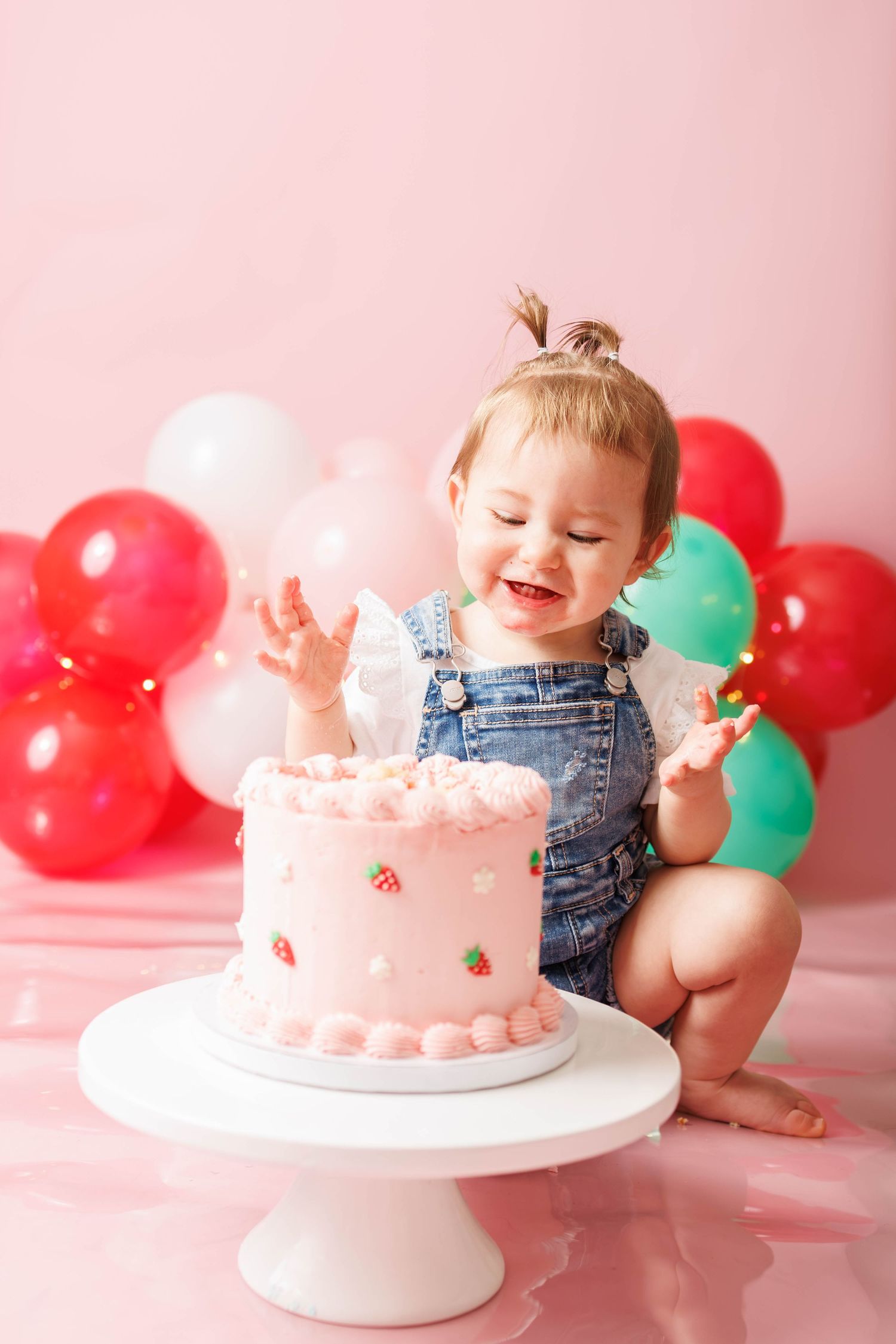 Happy toddler girl in denim overalls smashing her first birthday pink strawberry cake surrounded by colorful balloons.