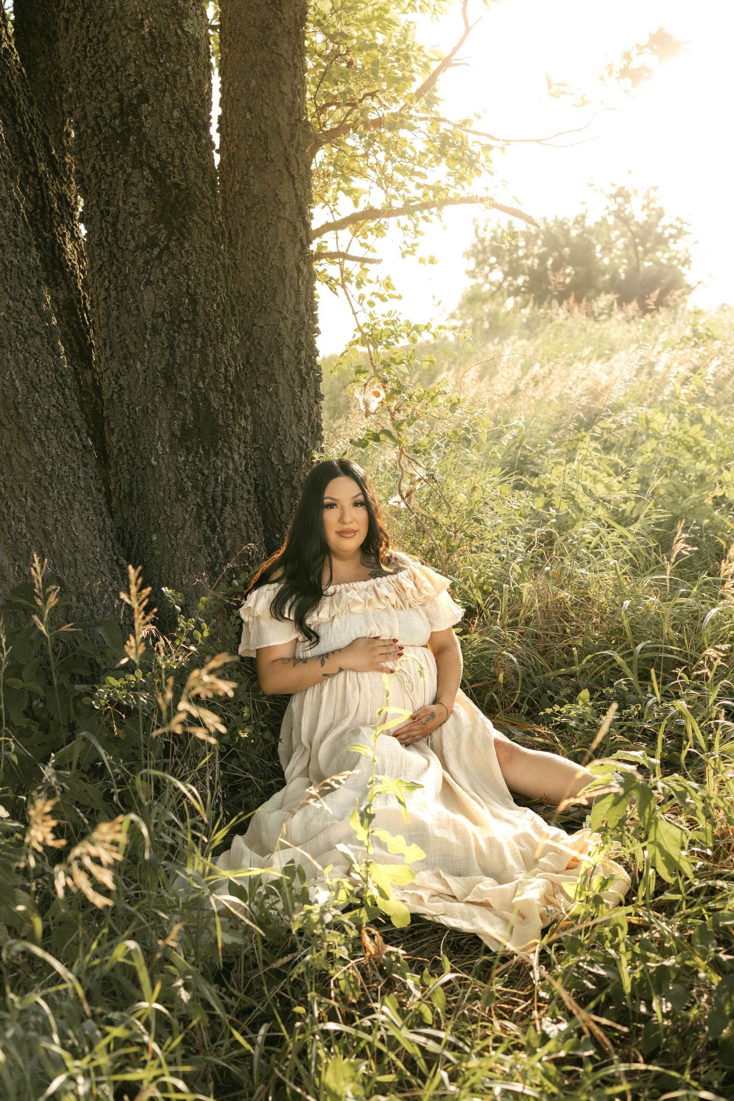 Pregnant woman in white dress sitting against a large tree in a sunlit meadow, cradling her baby bump.