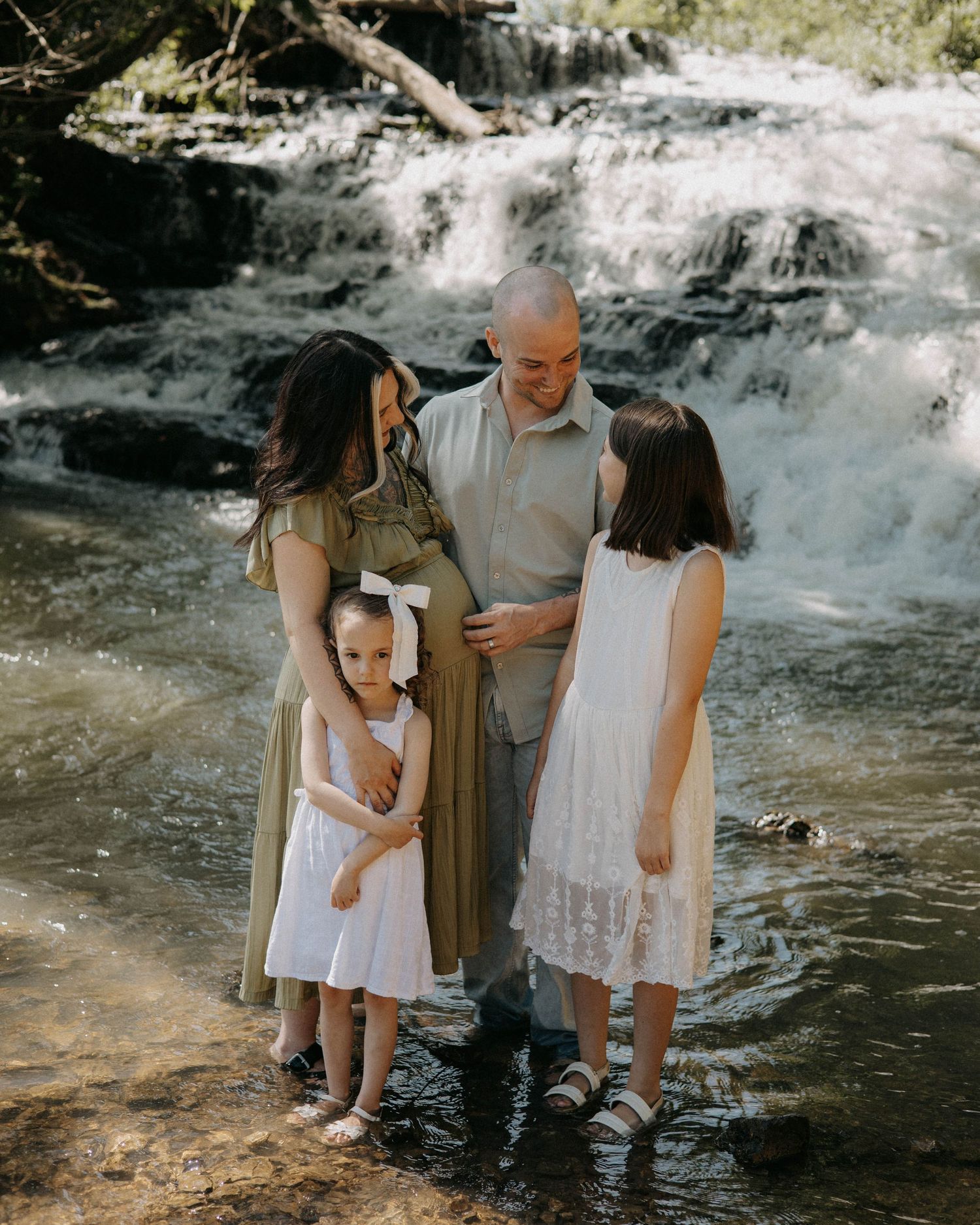 A family of four poses together in front of a cascading waterfall in nature wearing light colored dresses.