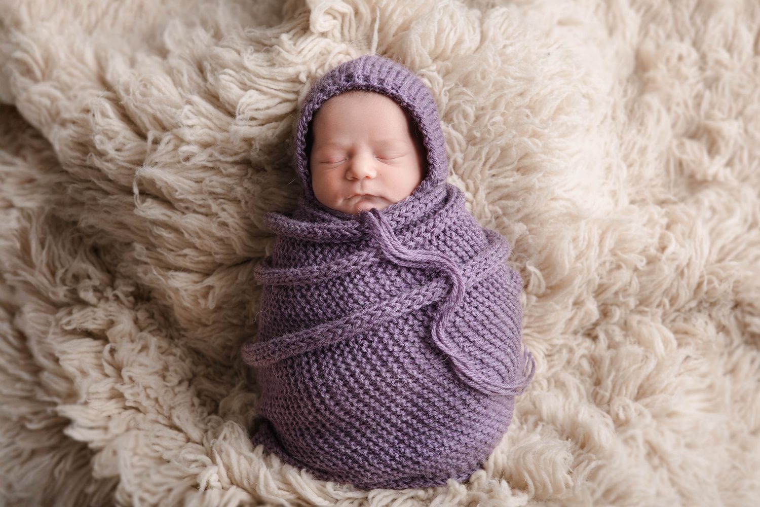 Sleeping newborn wrapped in purple knitted blanket on fluffy cream background.