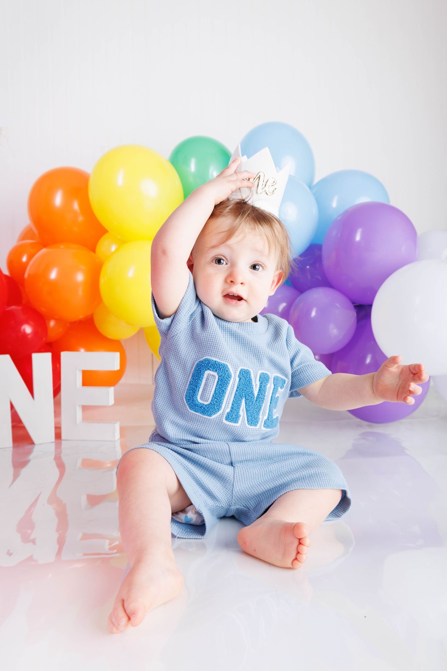 A baby in a blue outfit sits on a white backdrop surrounded by colorful rainbow balloons for a first birthday photo shoot.