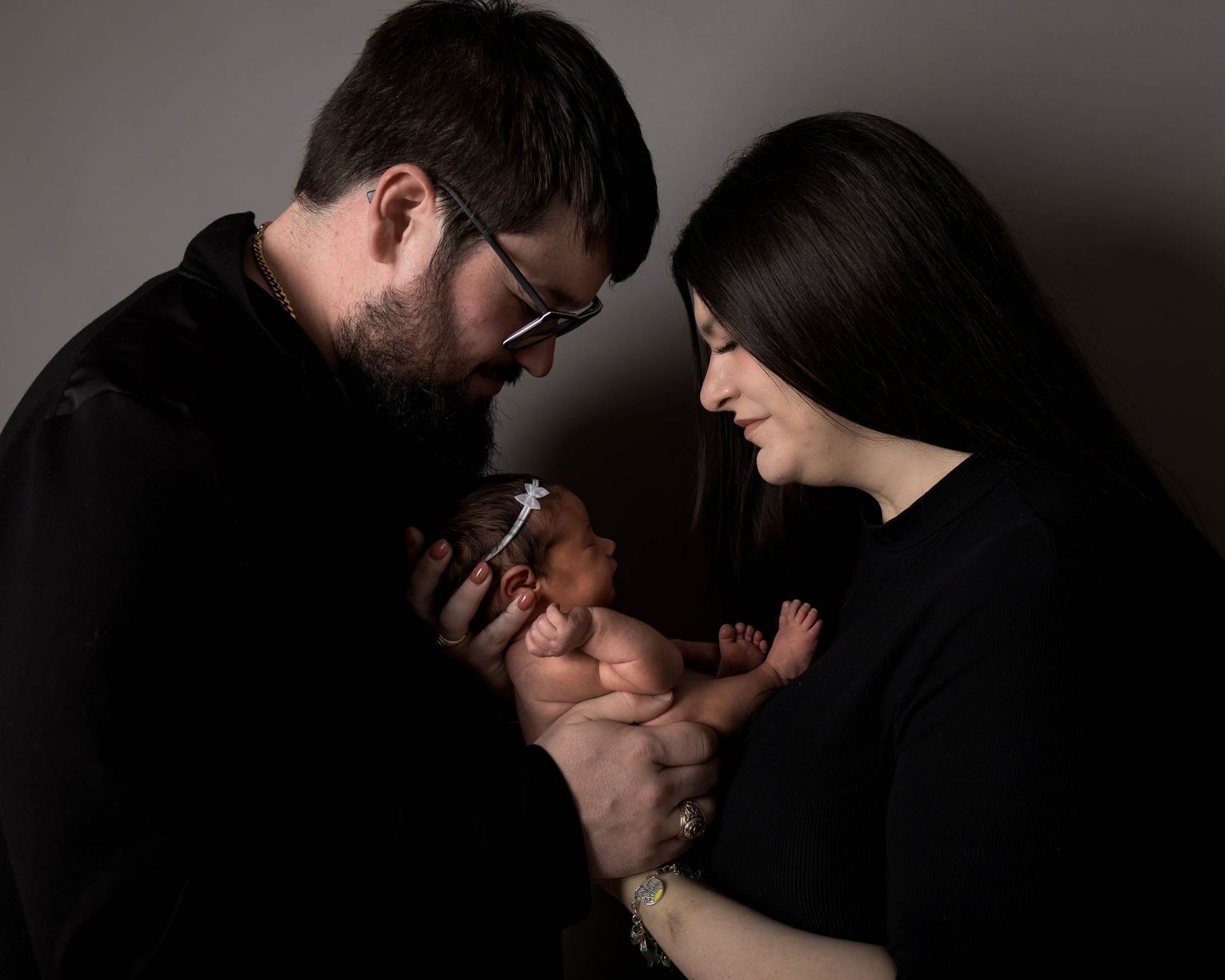 New parents in black outfits tenderly hold their newborn baby together against a gray studio background.