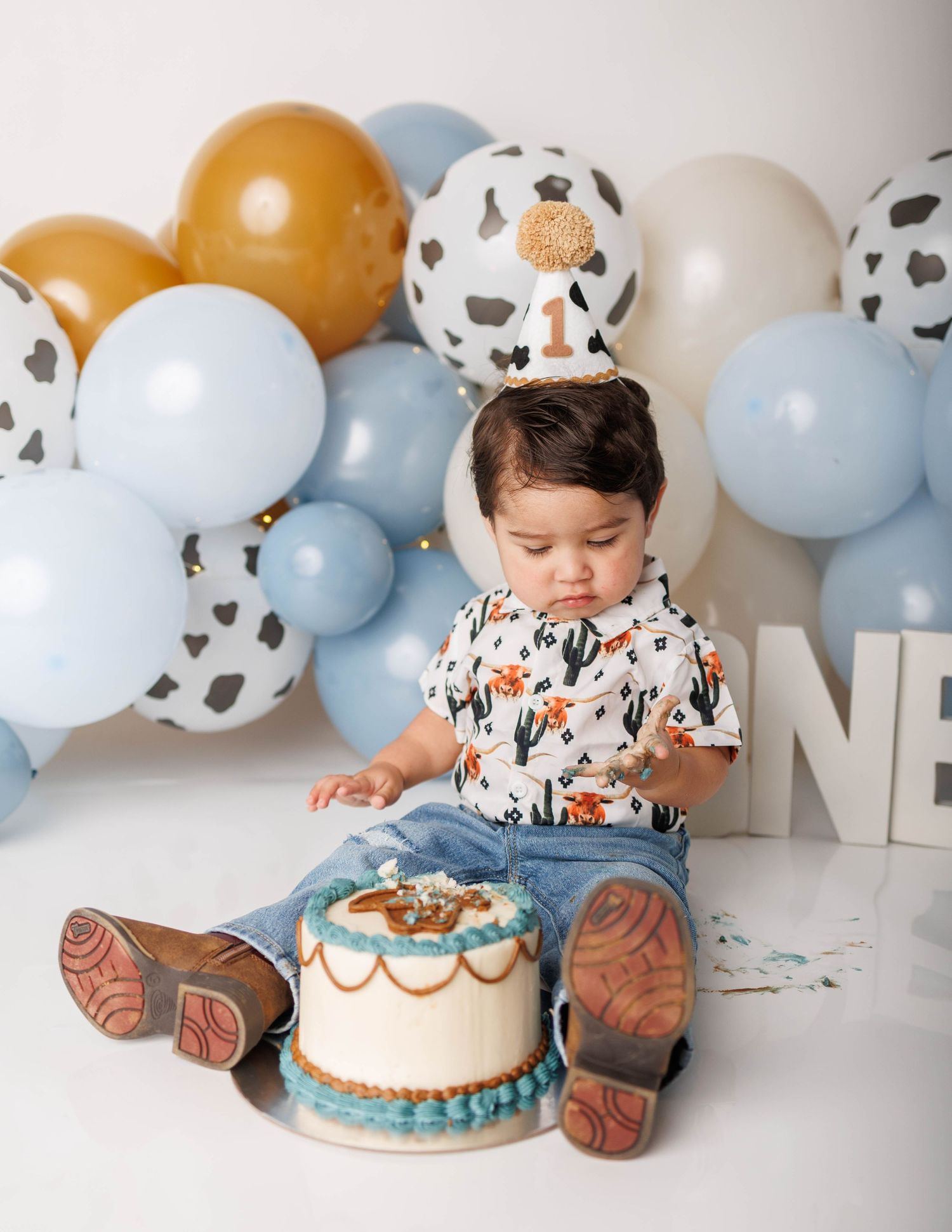 Baby boy in cowboy hat and boots smashes first birthday cake surrounded by blue and gold balloon garland.