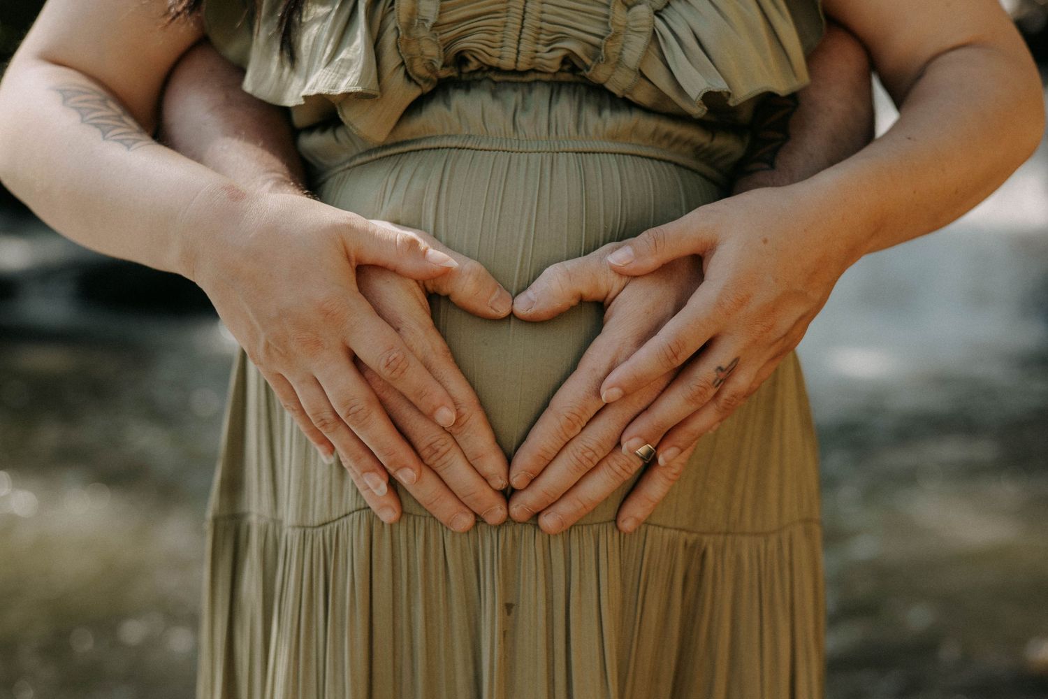 Close up shot of hands forming a heart shape over a pregnant belly in an olive green dress.