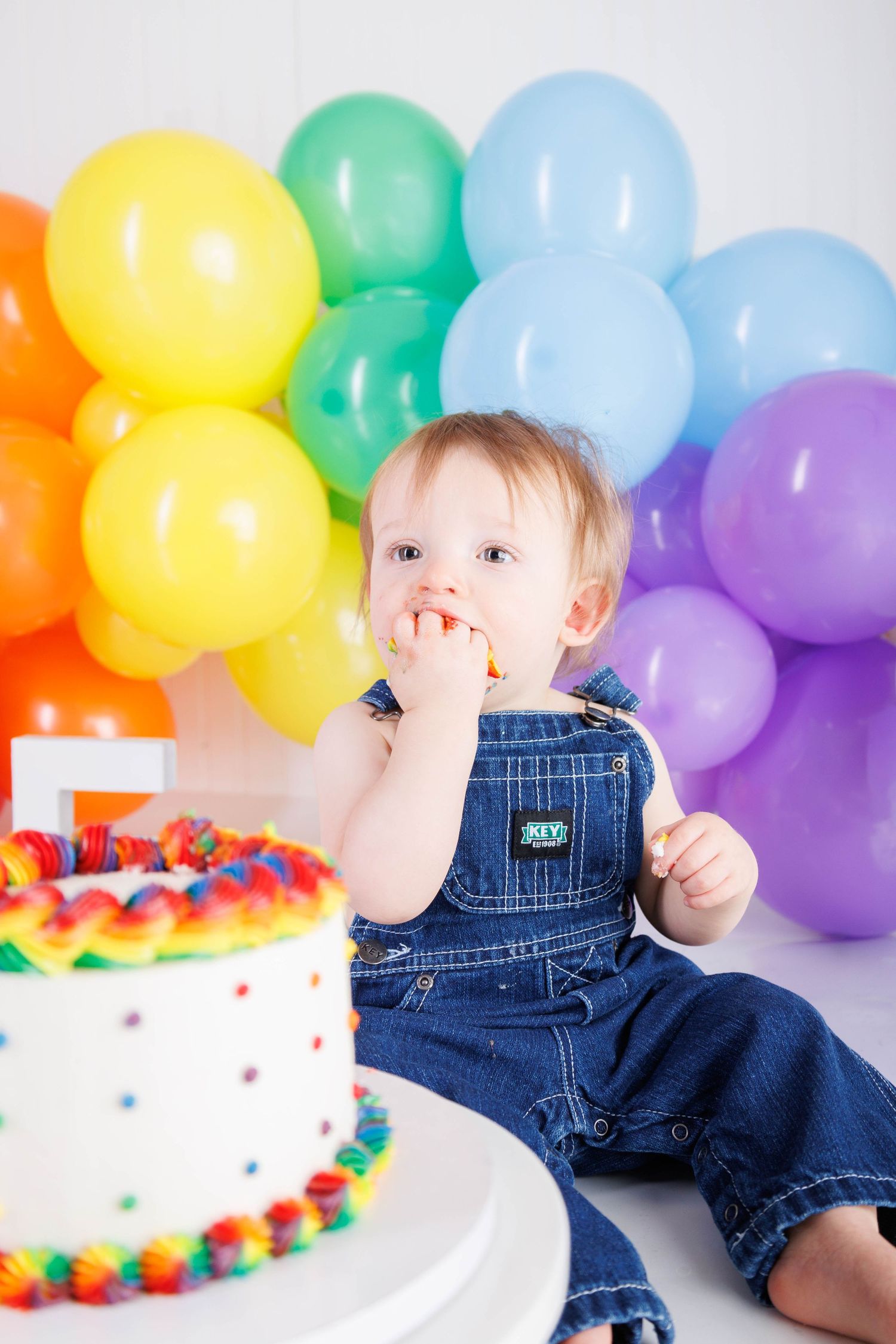 A series of photos showing a cake smash photoshoot with colorful balloons and a white frosted birthday cake.