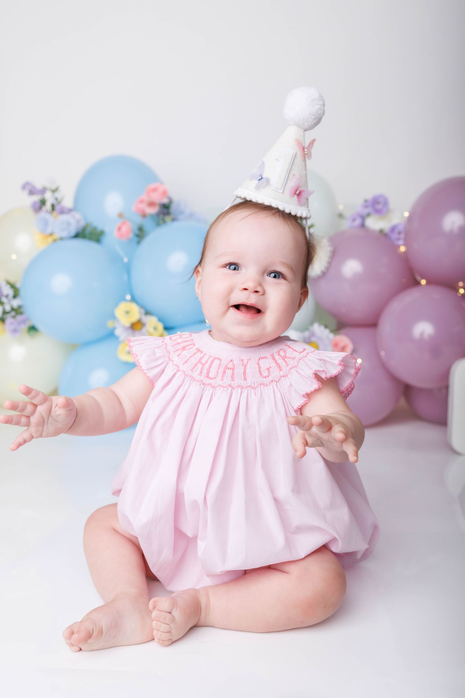 Happy baby girl in pink birthday dress and party hat sits surrounded by colorful pastel balloons.