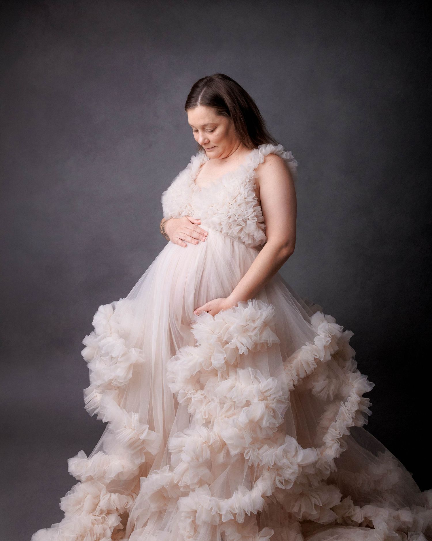 Pregnant woman in a flowing blush ruffle tulle gown cradling her baby bump in a professional maternity studio photo.