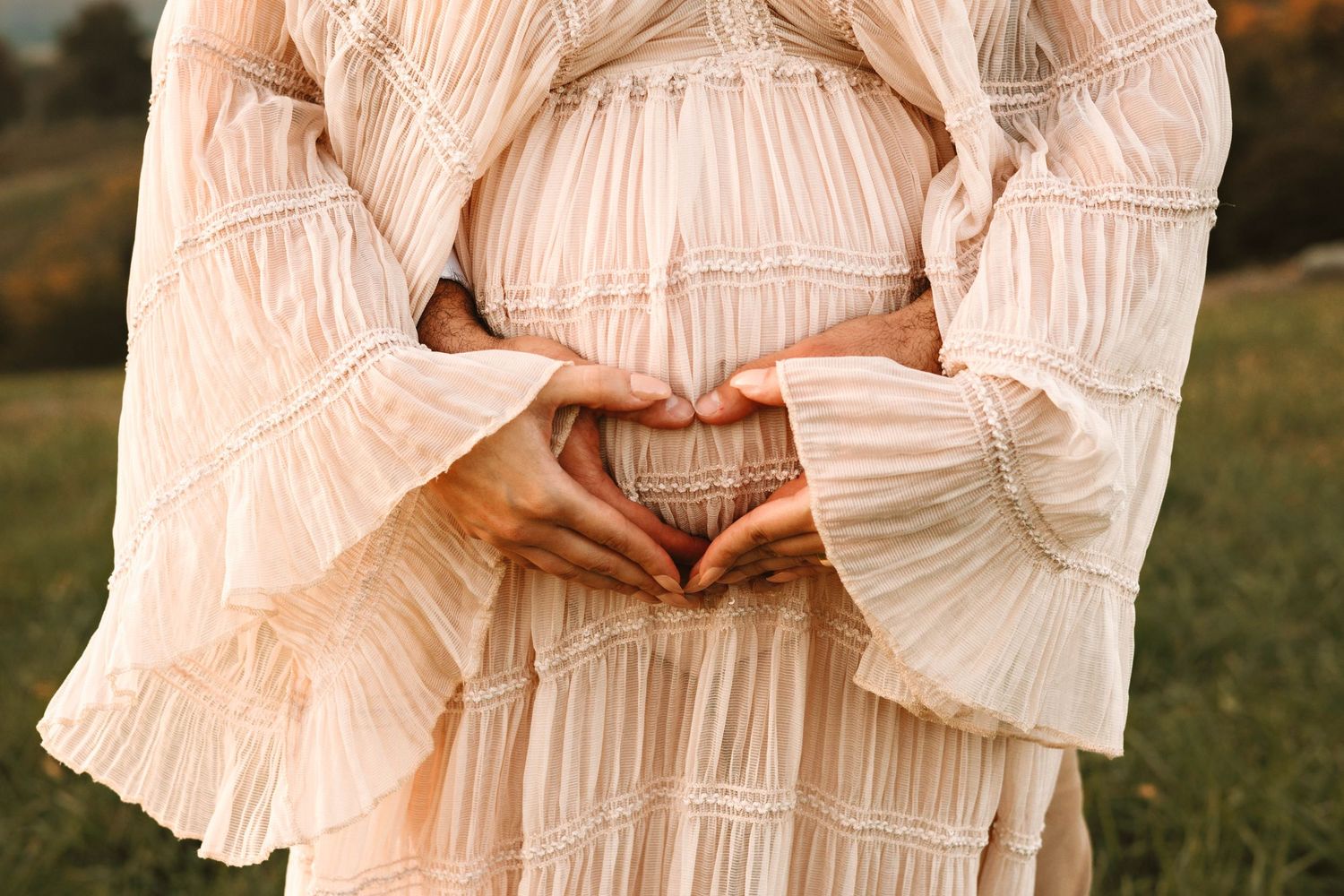 Pregnant woman in a boho peach dress forming a heart shape with her hands over her baby bump outdoors.