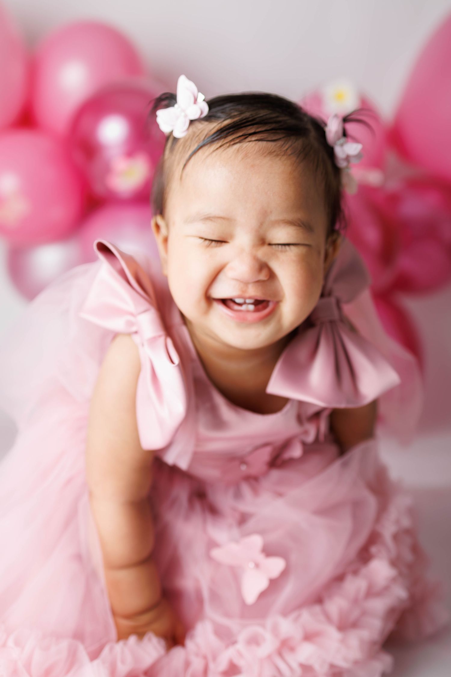 Adorable baby girl laughing joyfully in pink ruffle dress with butterfly hair clips and pink balloons backdrop.