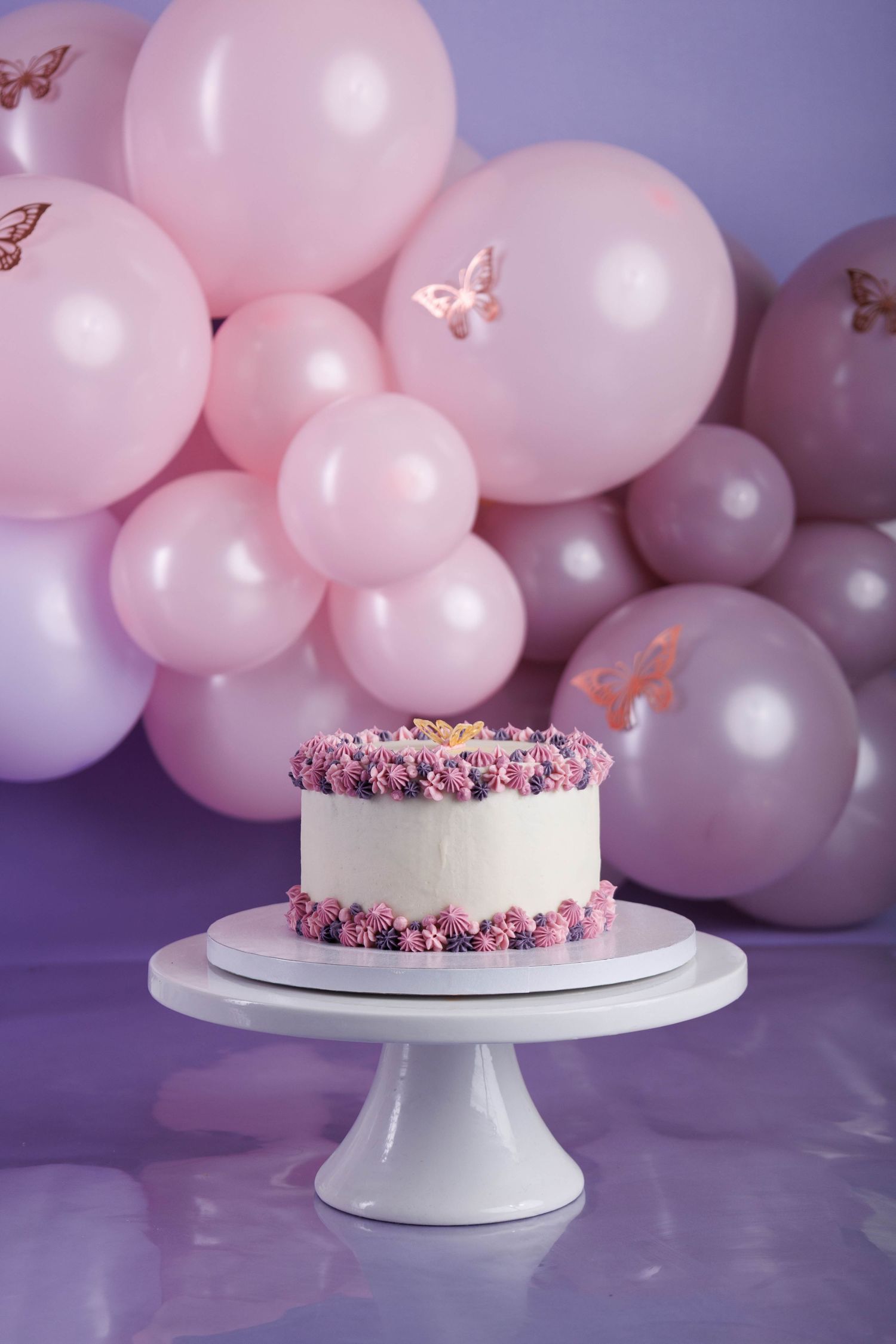 White frosted cake with pink floral decorations on cake stand against purple backdrop with pink balloon garland.