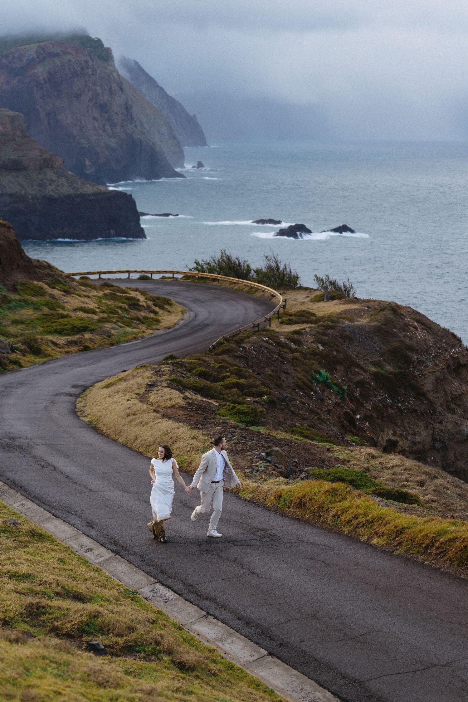 Bride and groom holding hands walking along a winding coastal road with dramatic cliffs and ocean views in Madeira.