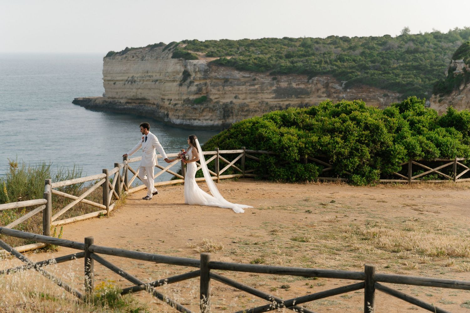 Newlyweds holding hands strolling along rustic fenced coastal path with dramatic cliffs and turquoise sea.