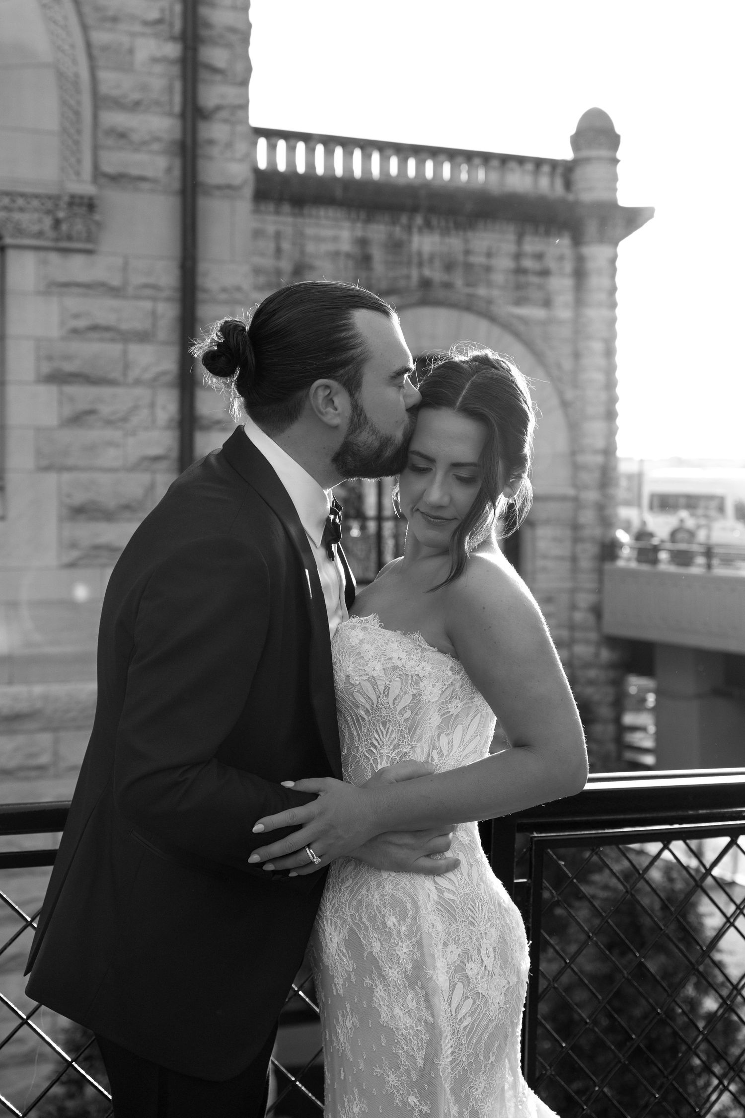 Black and white photo of groom kissing bride on cheek while embracing outdoors near ornate stone building.