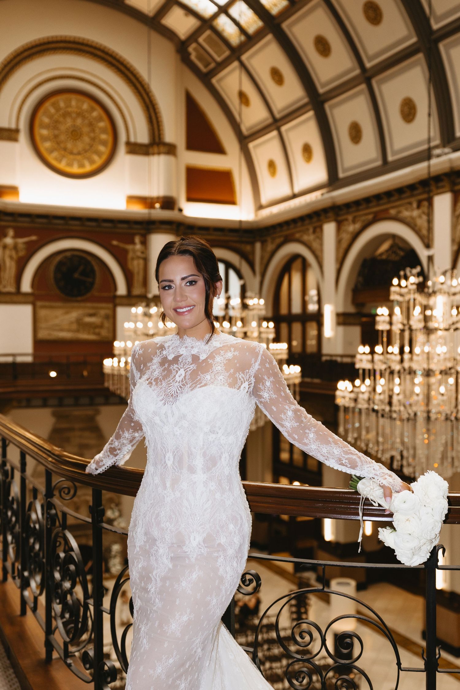 Bride in long-sleeve lace wedding gown holding white bouquet on ornate staircase in elegant ballroom venue.