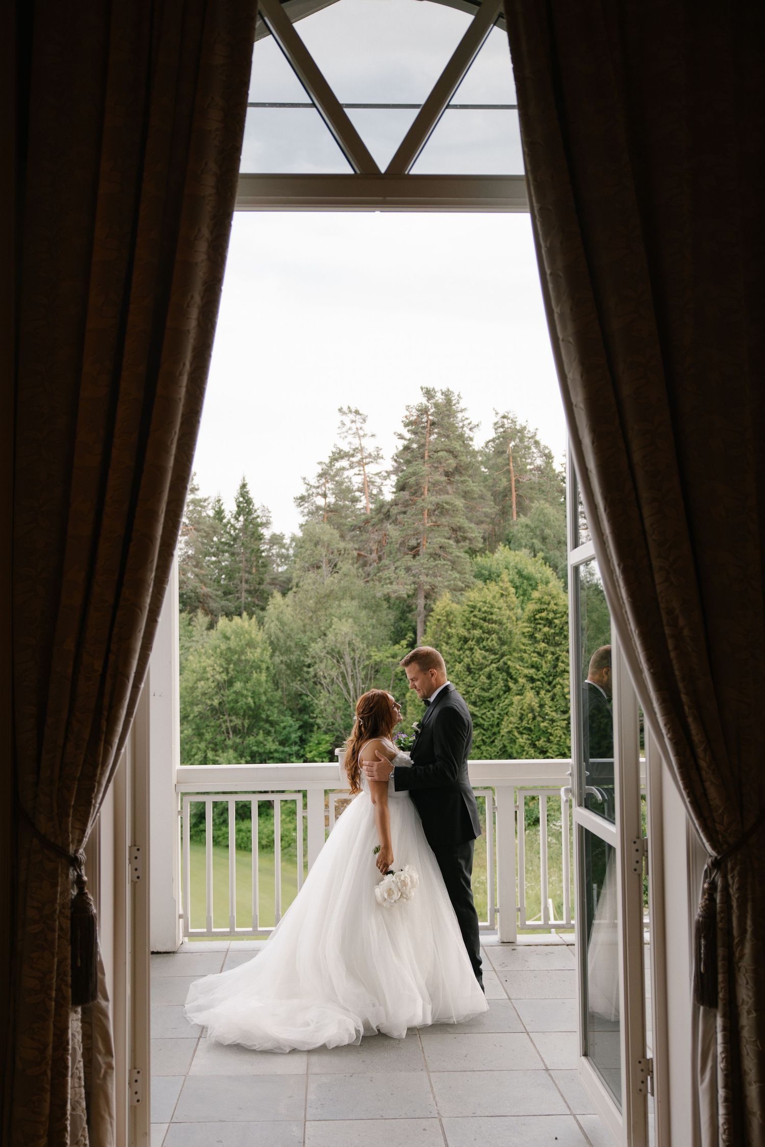 Bride and groom standing on a balcony surrounded by lush green trees, viewed through elegant curtained doorway.