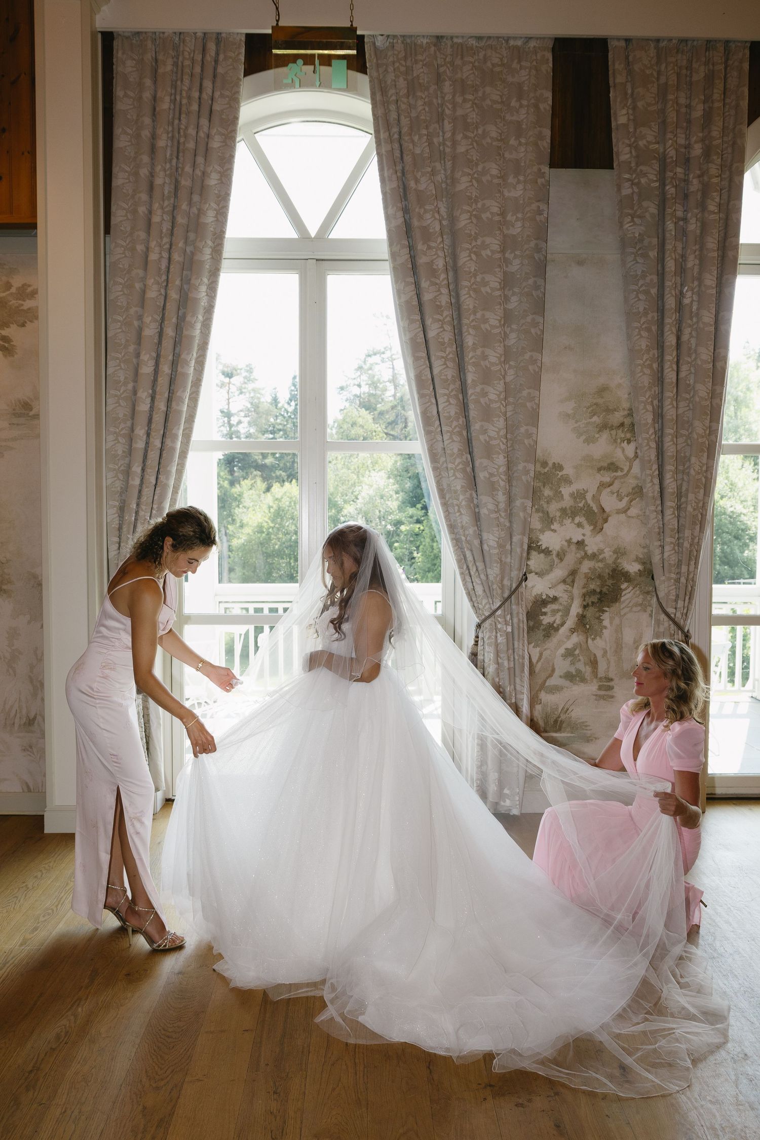 Bride in white ballgown with veil being assisted by two bridesmaids in pink near large window.