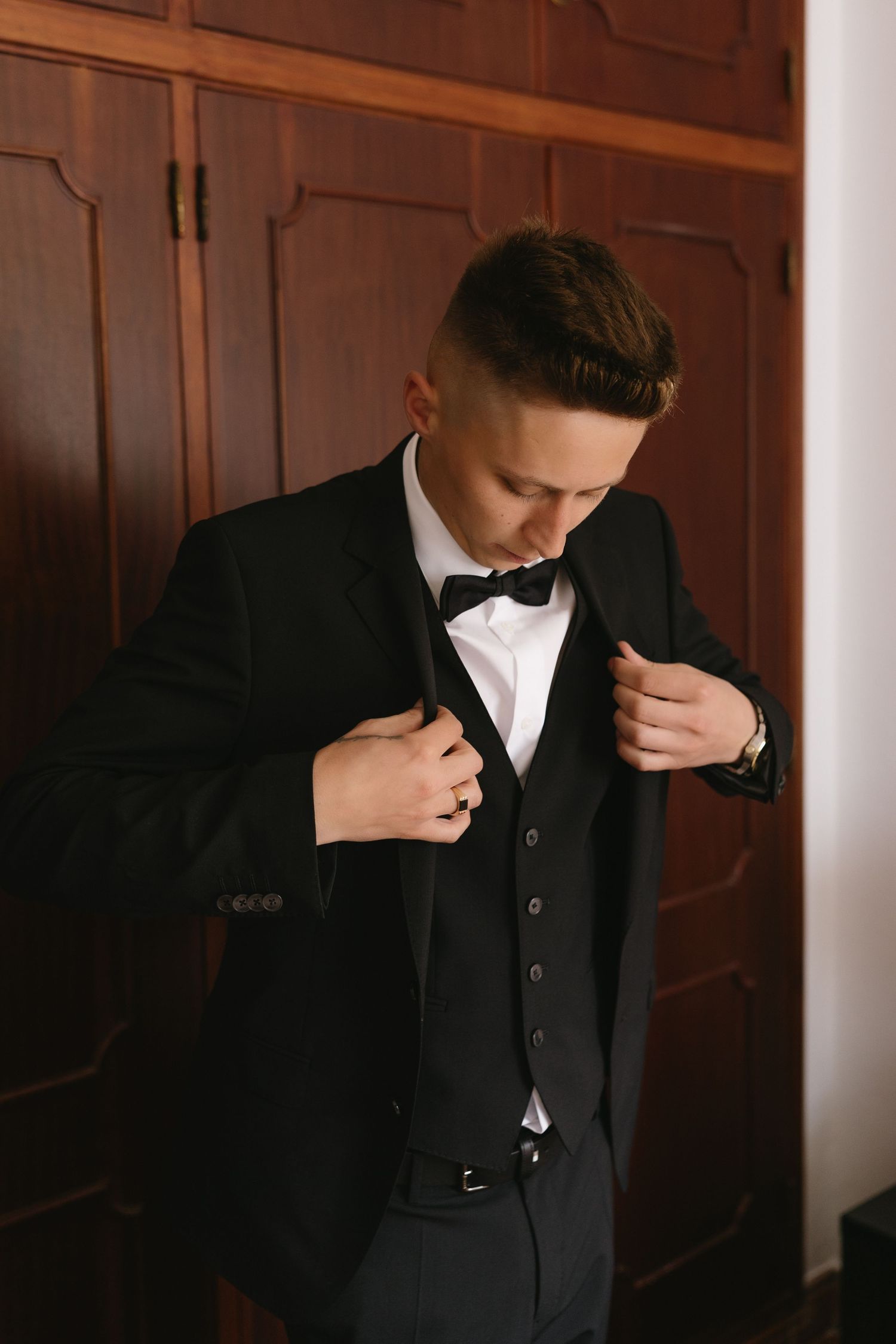 Groom in black tuxedo with bow tie adjusting his jacket lapels before wedding ceremony.