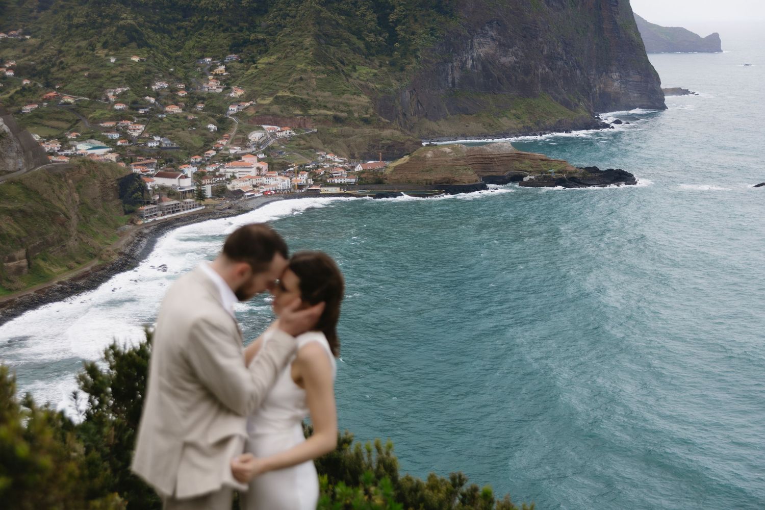 A couple shares a romantic moment on a cliff overlooking a turquoise bay and coastal village in Madeira.