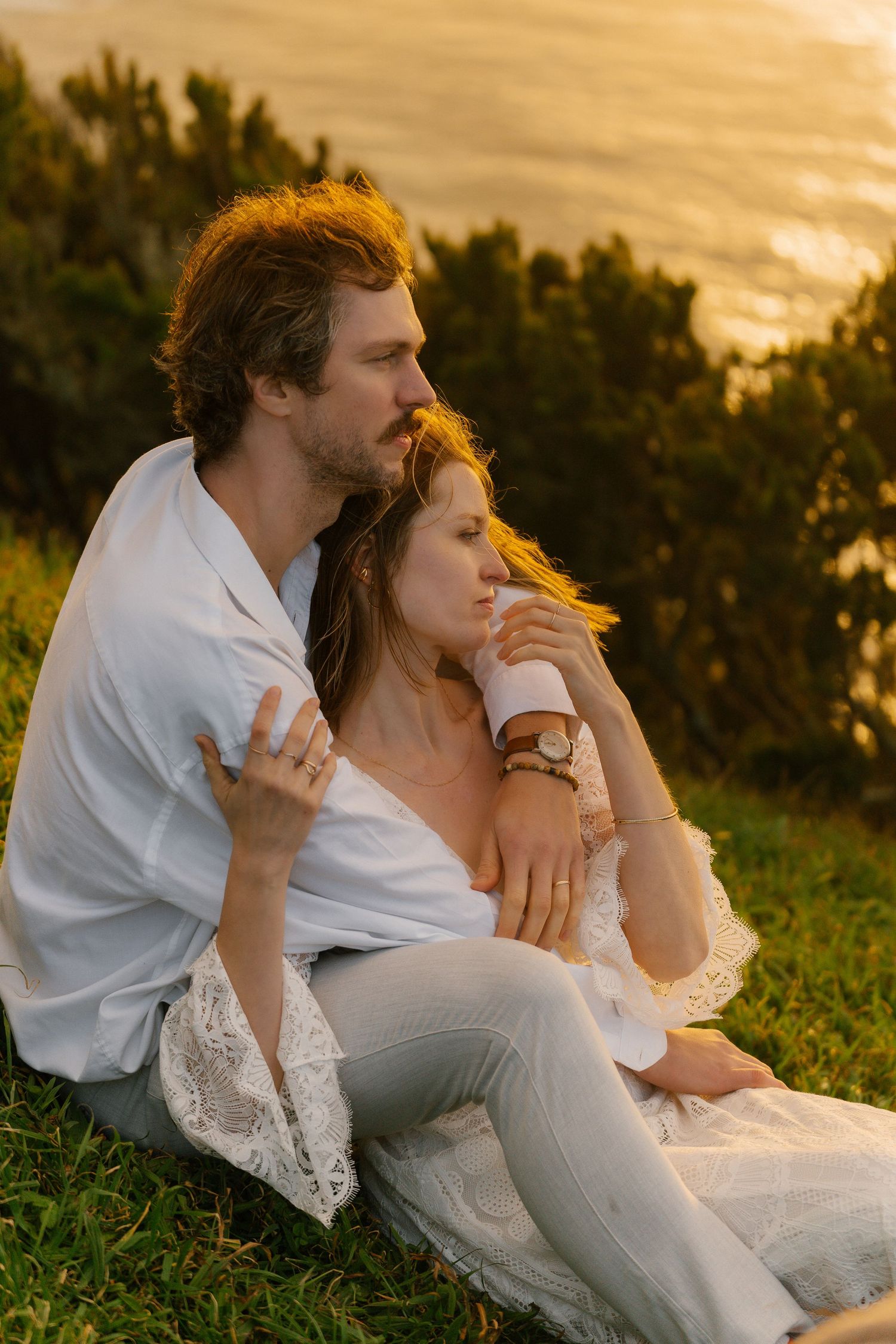 Couple sitting together on a grassy hillside at golden hour, groom embracing bride with ocean view behind.