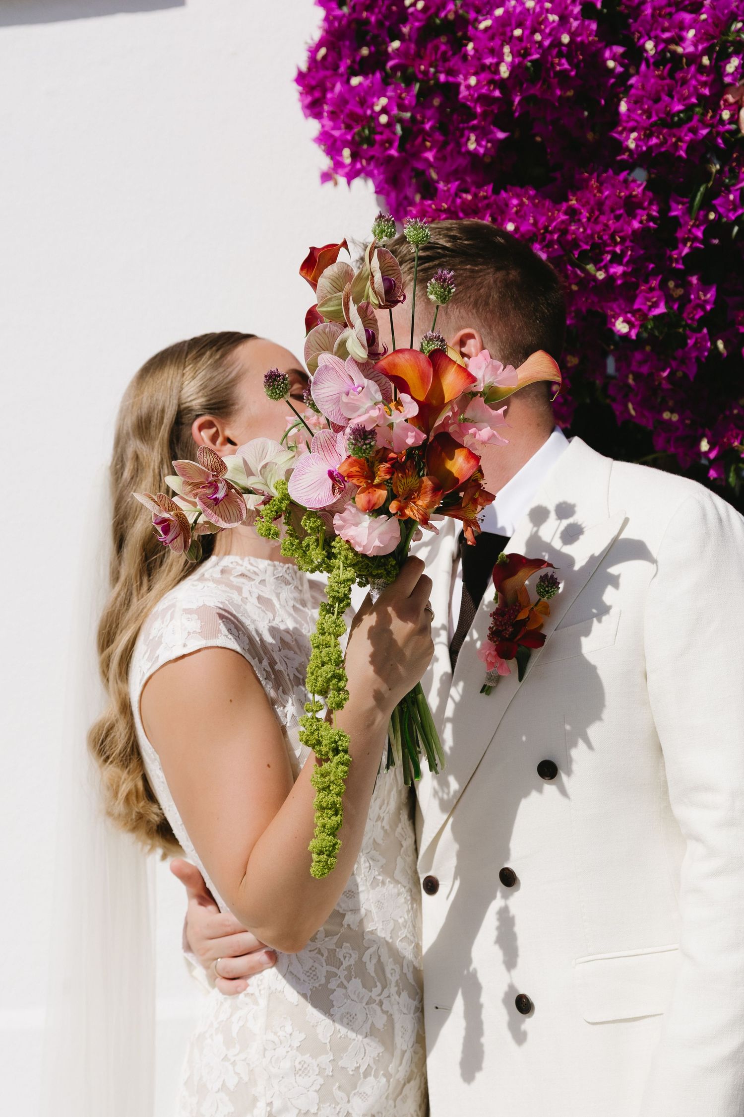 Bride and groom kiss behind vibrant tropical bouquet with purple bougainvillea blooming in background.