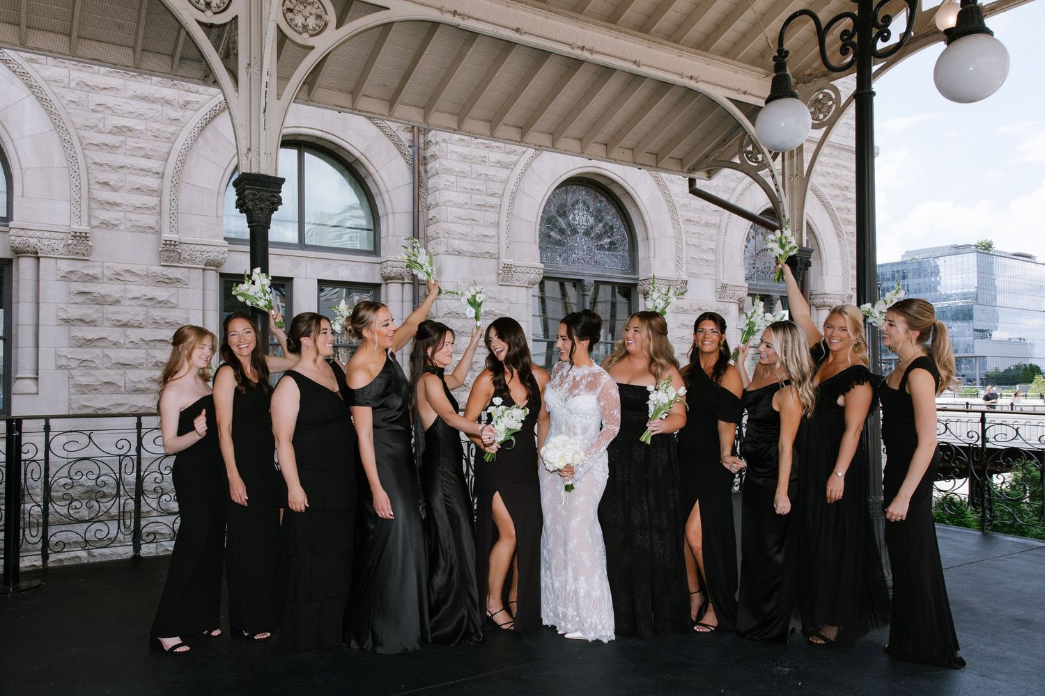 Bride in white gown with bridesmaids in black dresses raising bouquets outside elegant stone building.