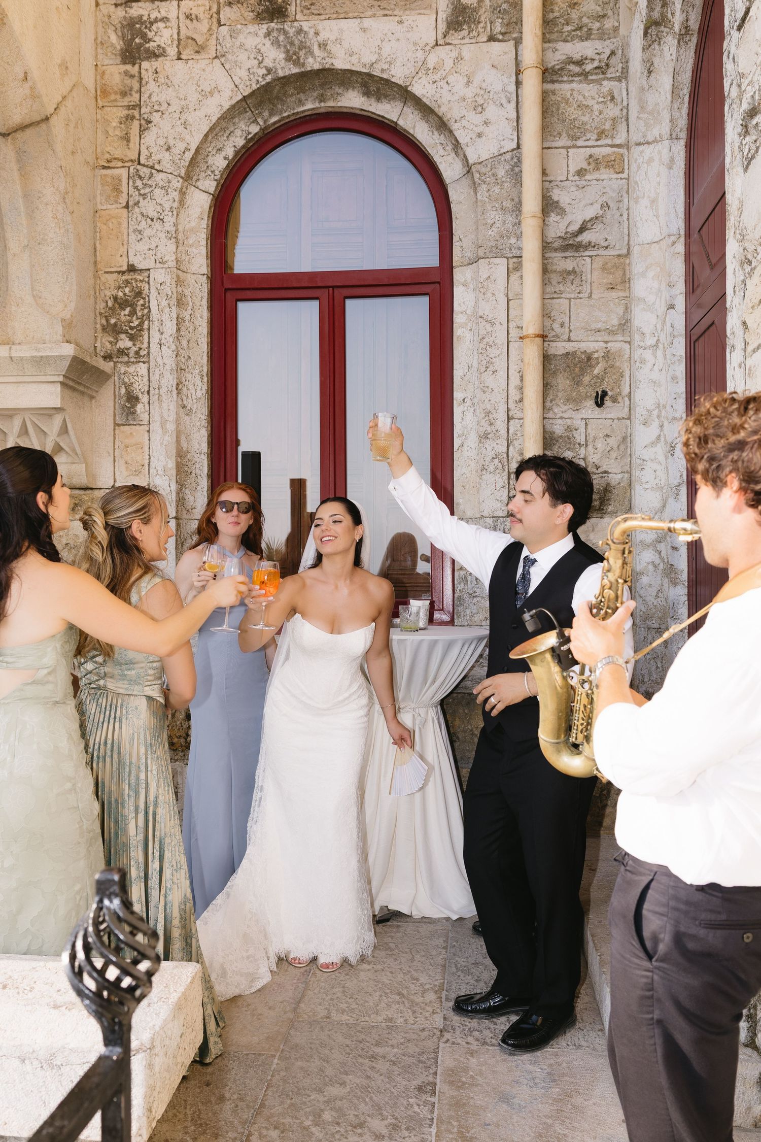Bride and groom toast with wedding party beside saxophonist at elegant stone venue.