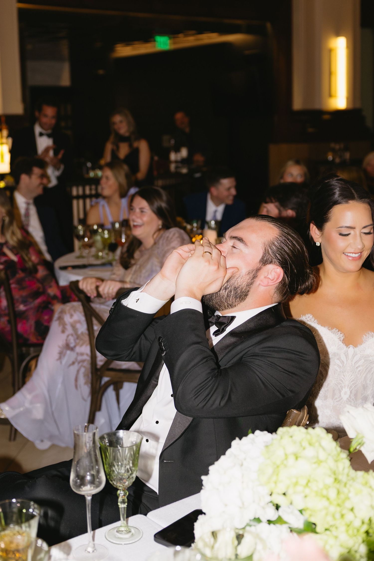 Groom in tuxedo laughing and drinking at wedding reception head table with smiling bride in white gown beside him.