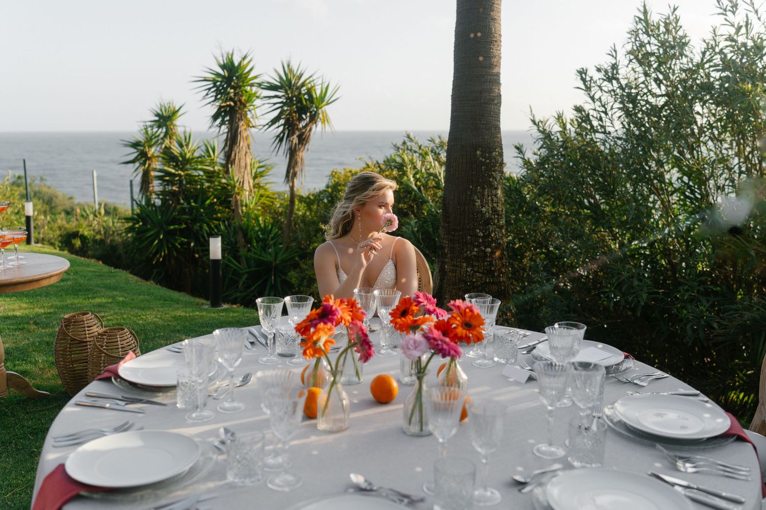 Blonde woman seated at elegantly set outdoor dining table with colorful flowers, overlooking ocean and tropical garden.
