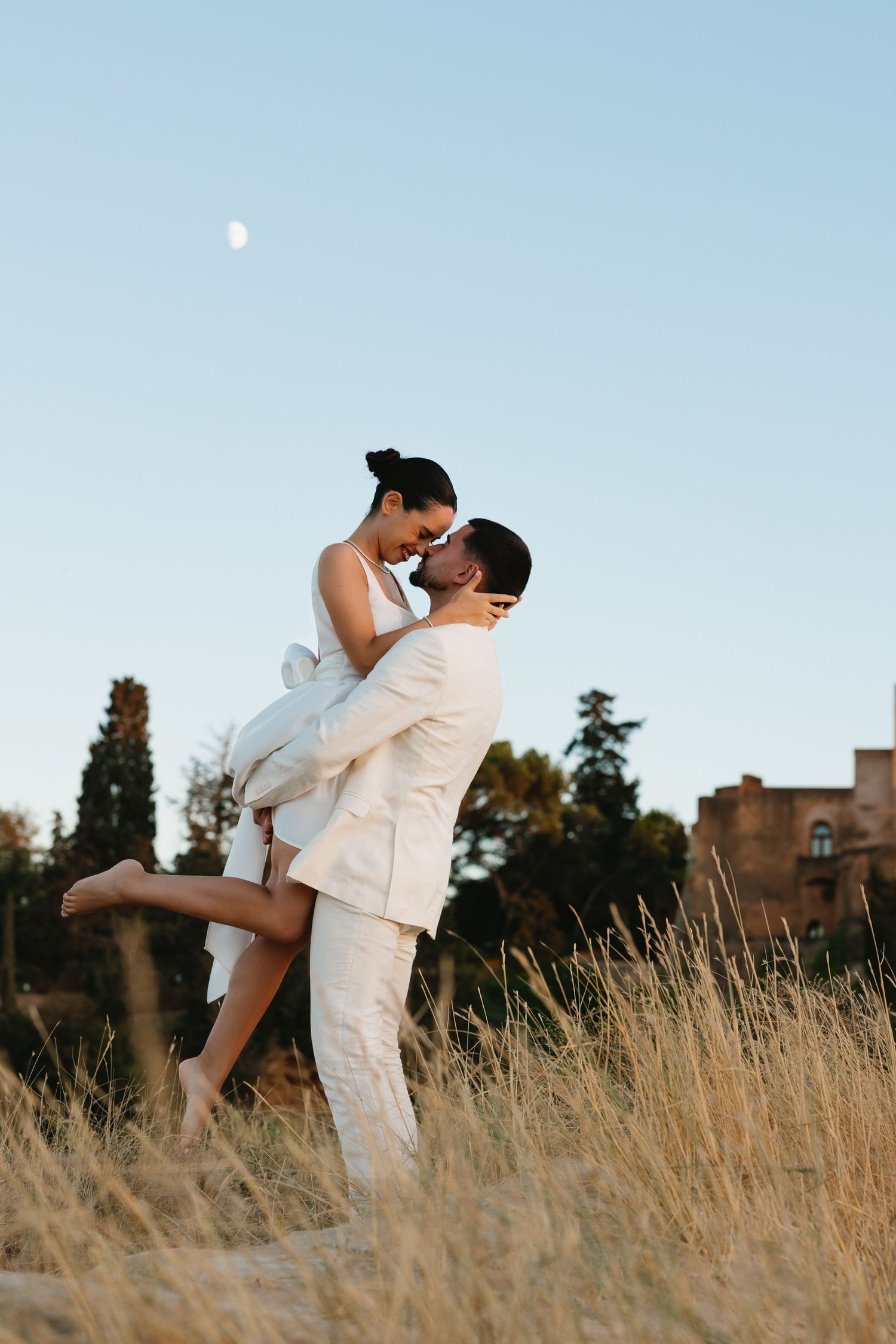 Groom lifts bride in white dress for a kiss in golden field under moonlit sky.