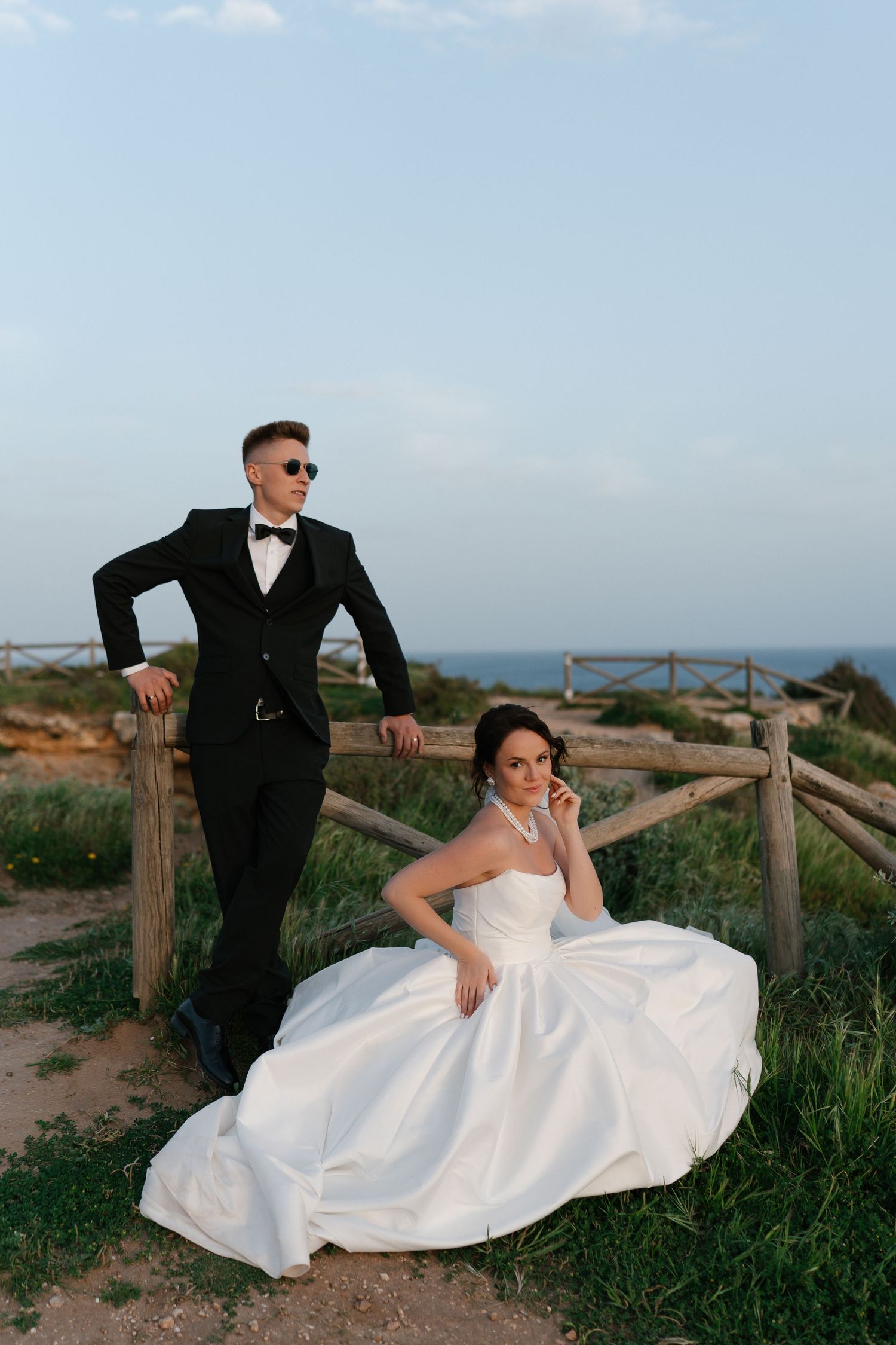Groom in black tuxedo leans on wooden fence while bride in white ballgown sits on grass by the sea.