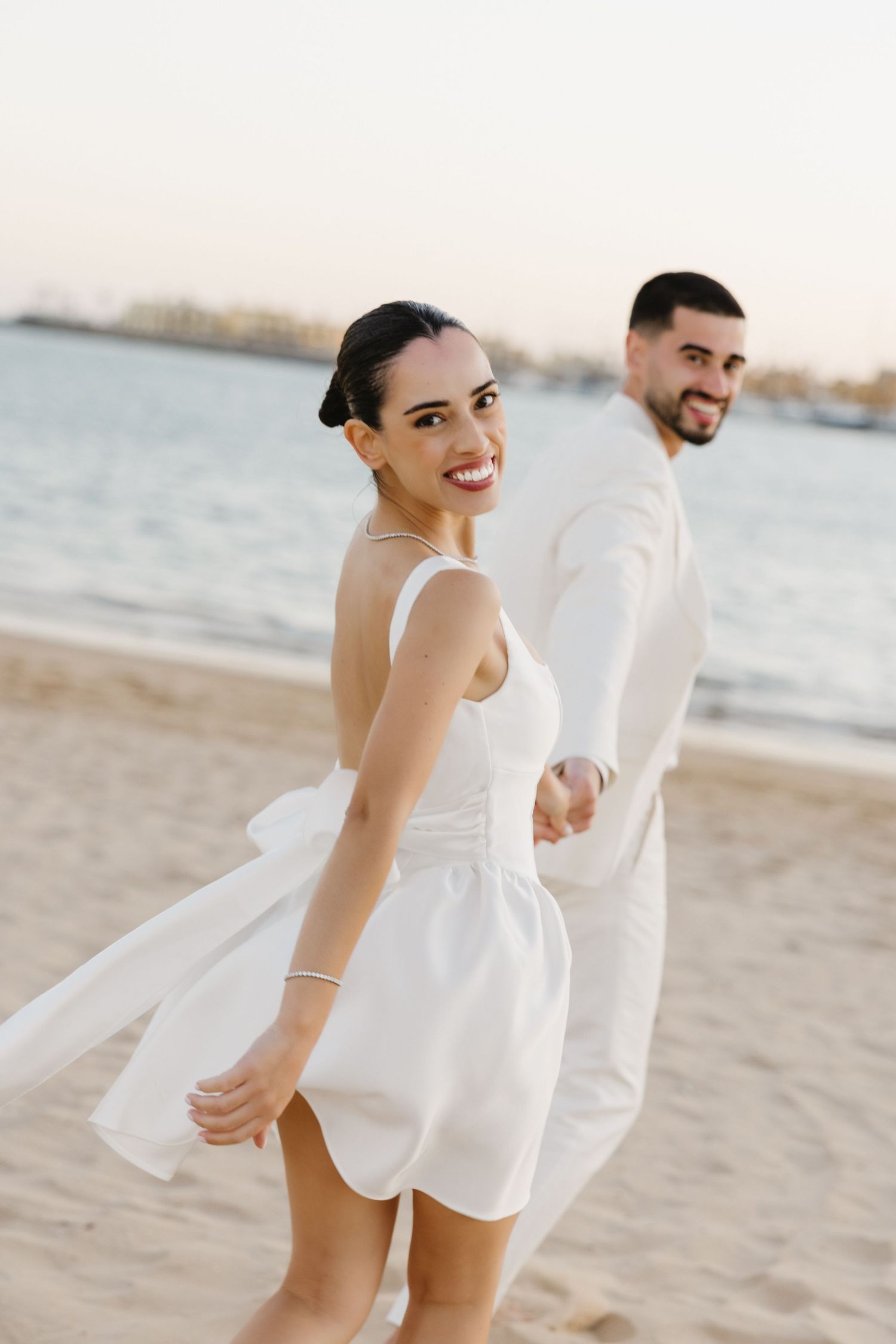 Joyful bride in white dress looks back smiling while holding groom's hand on sandy beach.
