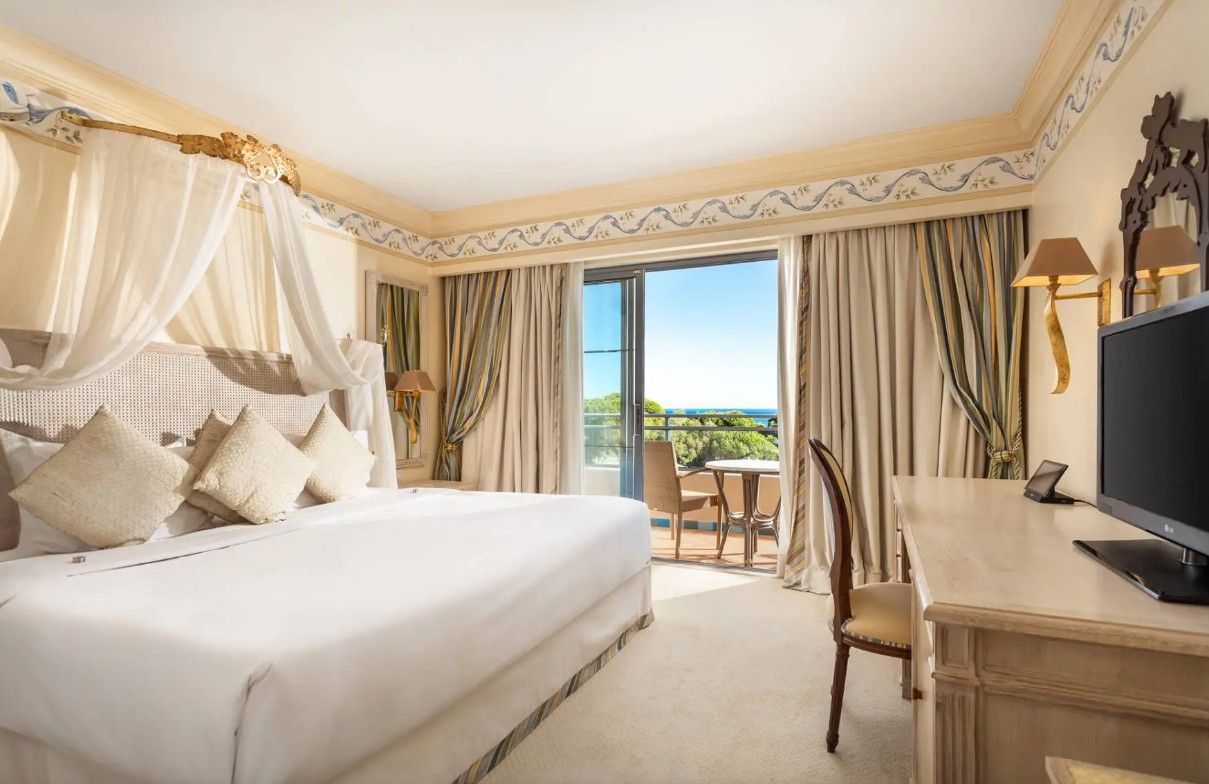 Luxurious hotel room with canopy bed, elegant decor, and balcony overlooking lush greenery.