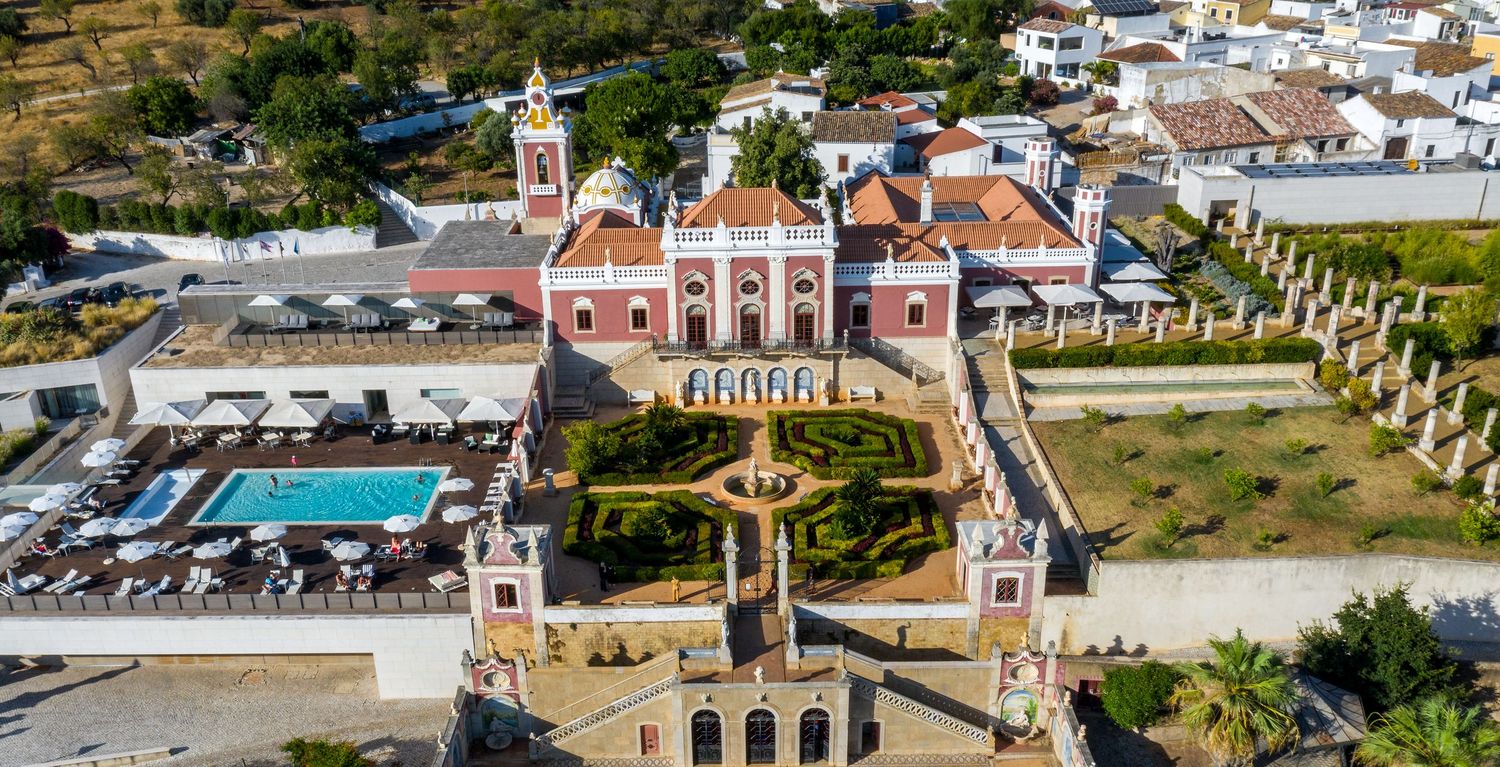 Aerial view of a grand pink baroque palace with formal gardens, pool, and surrounding historic village.