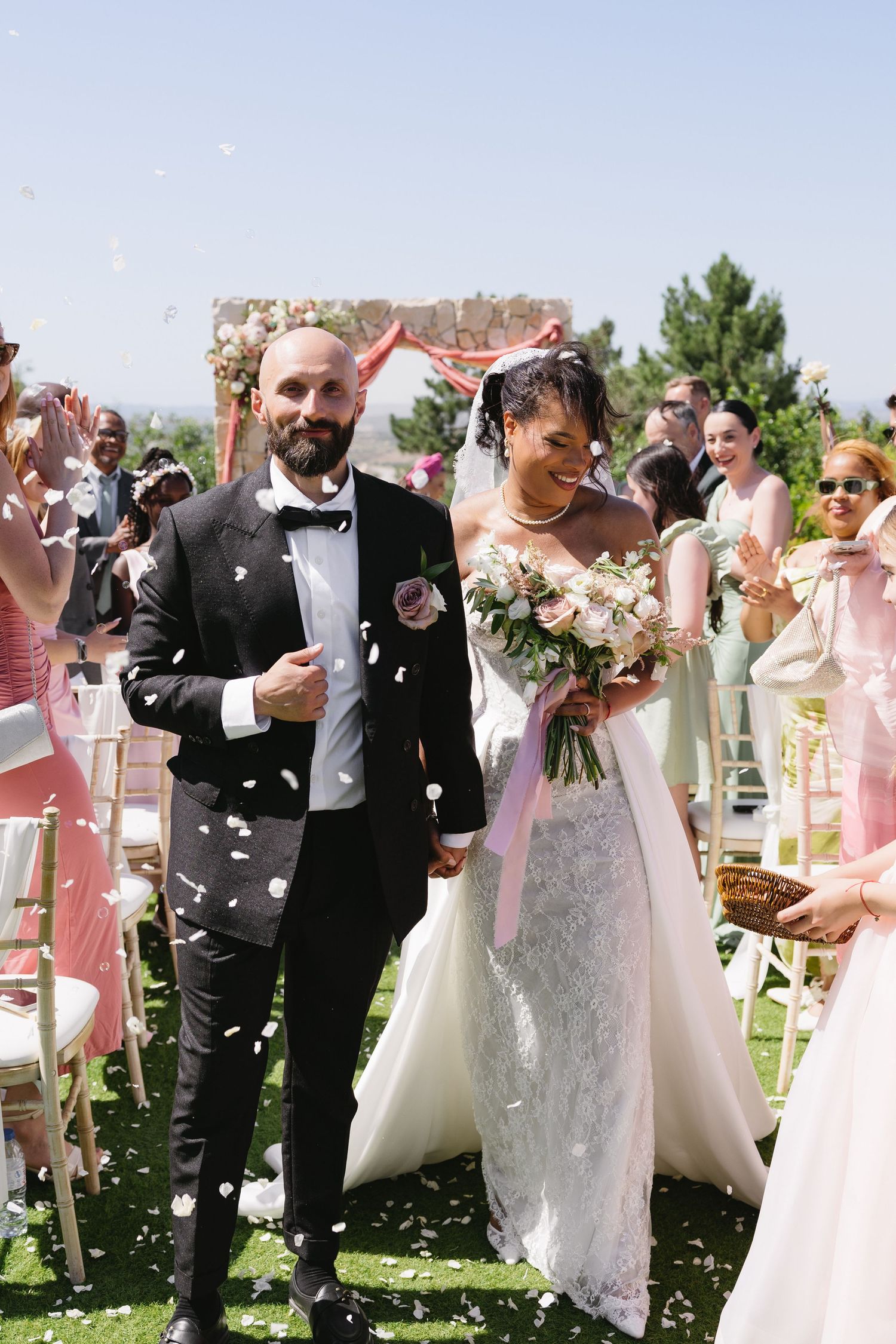 Smiling newlyweds walk down aisle as guests shower them with white flower petals at outdoor wedding ceremony.