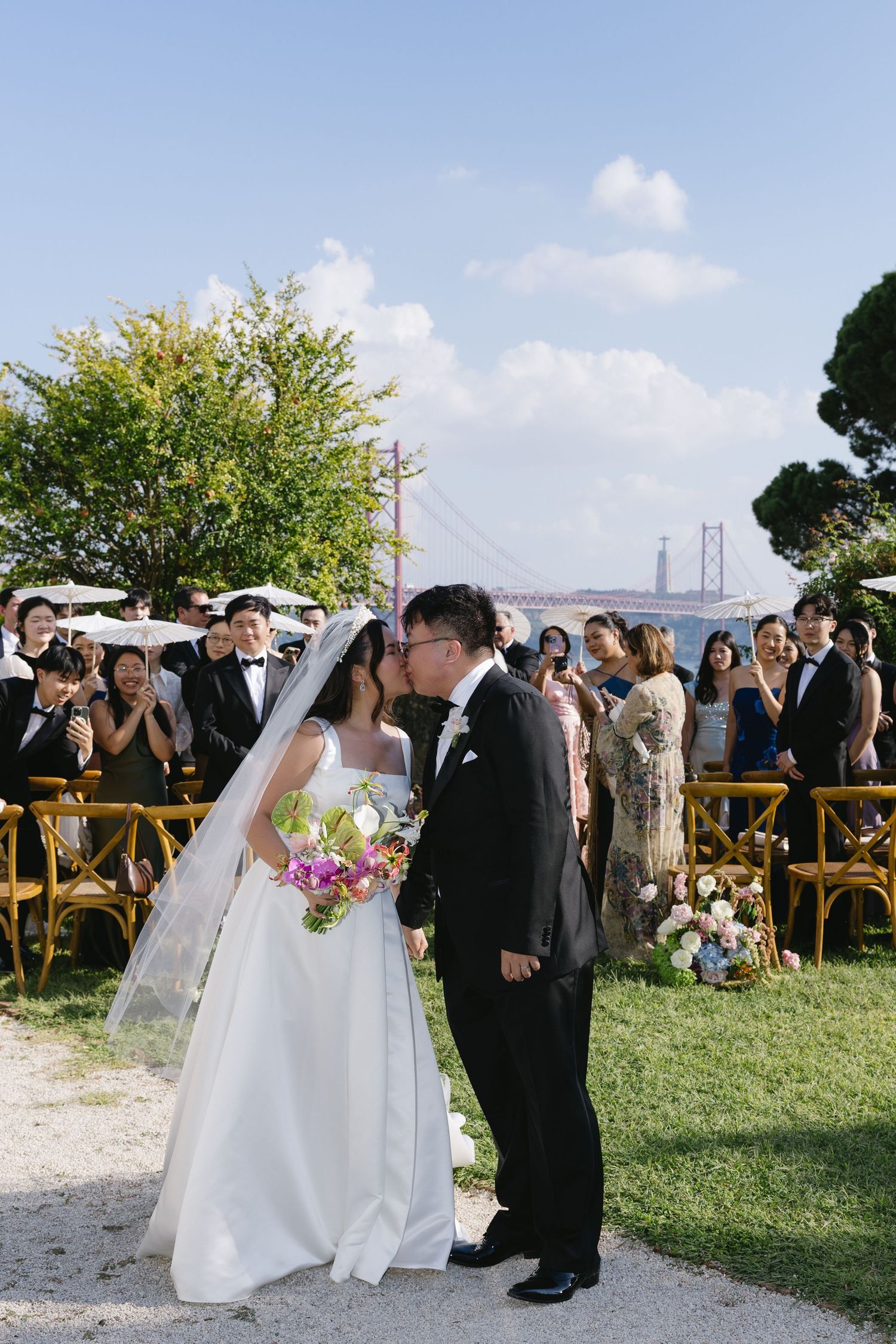 Bride and groom share a kiss at outdoor wedding ceremony with golden bridge and blue sky backdrop.