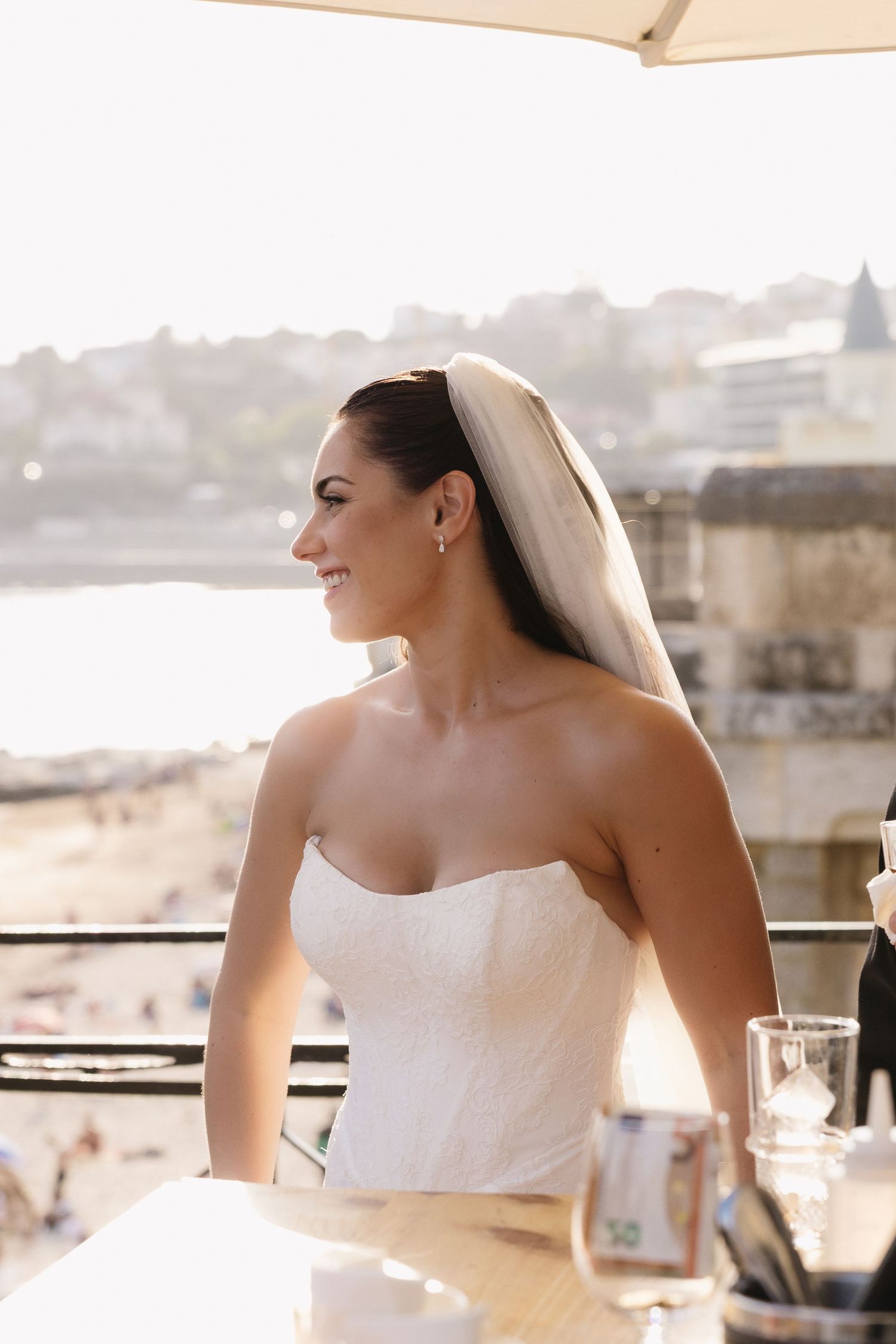 Smiling bride in strapless white gown and veil seated at a sunlit coastal venue with ocean views.