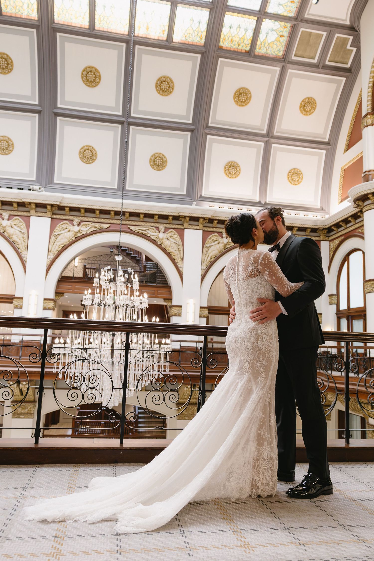 Bride and groom embrace in elegant ballroom with ornate ceiling, chandeliers, and intricate architectural details.