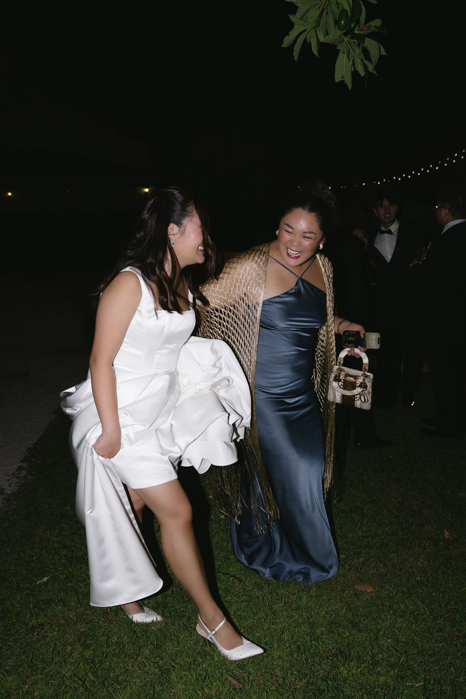 Two women in elegant formal attire laugh together outdoors at night during a wedding reception.