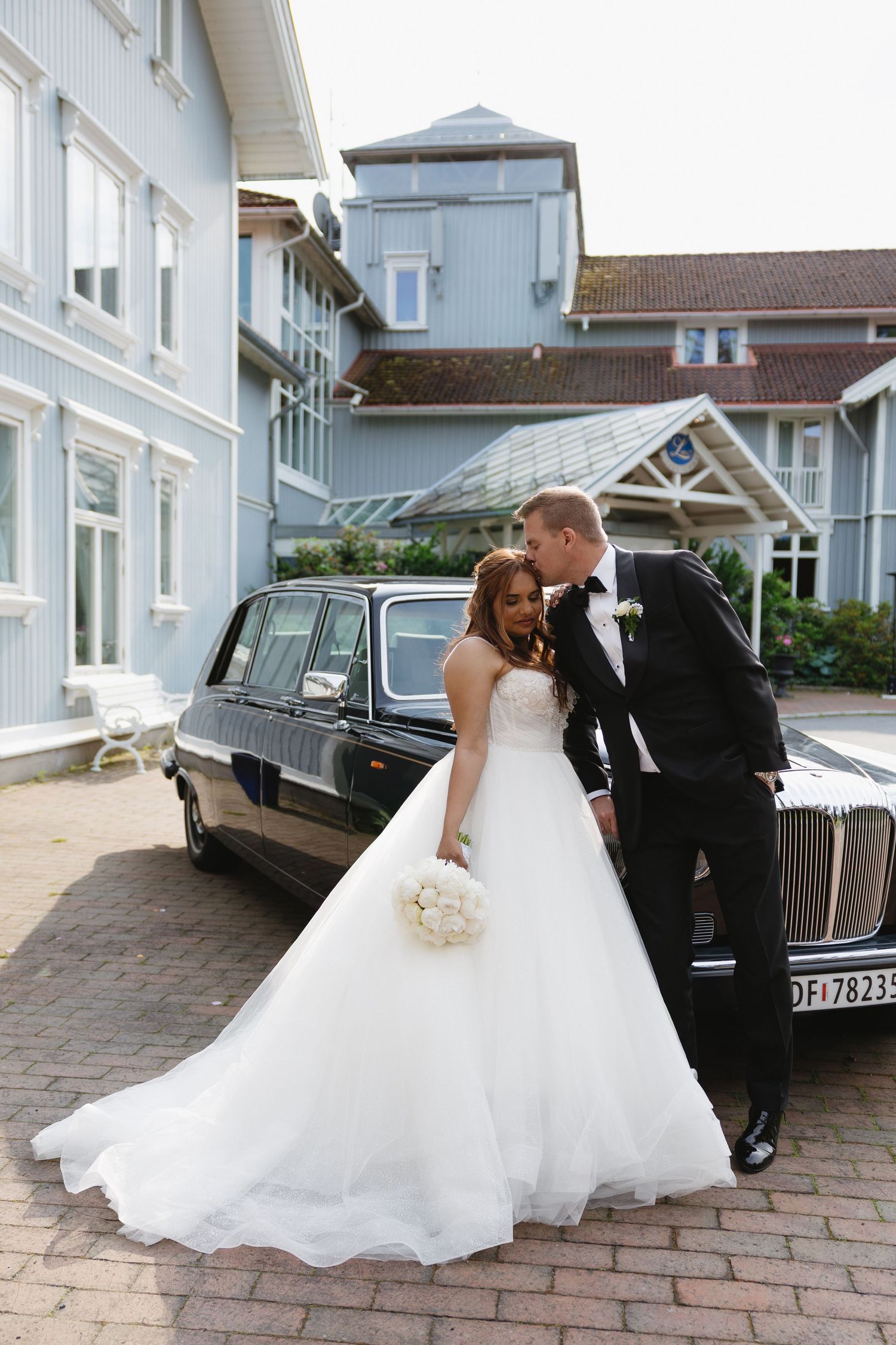 Bride in white ball gown and groom in tuxedo pose beside a black vintage limousine outside an elegant blue venue.